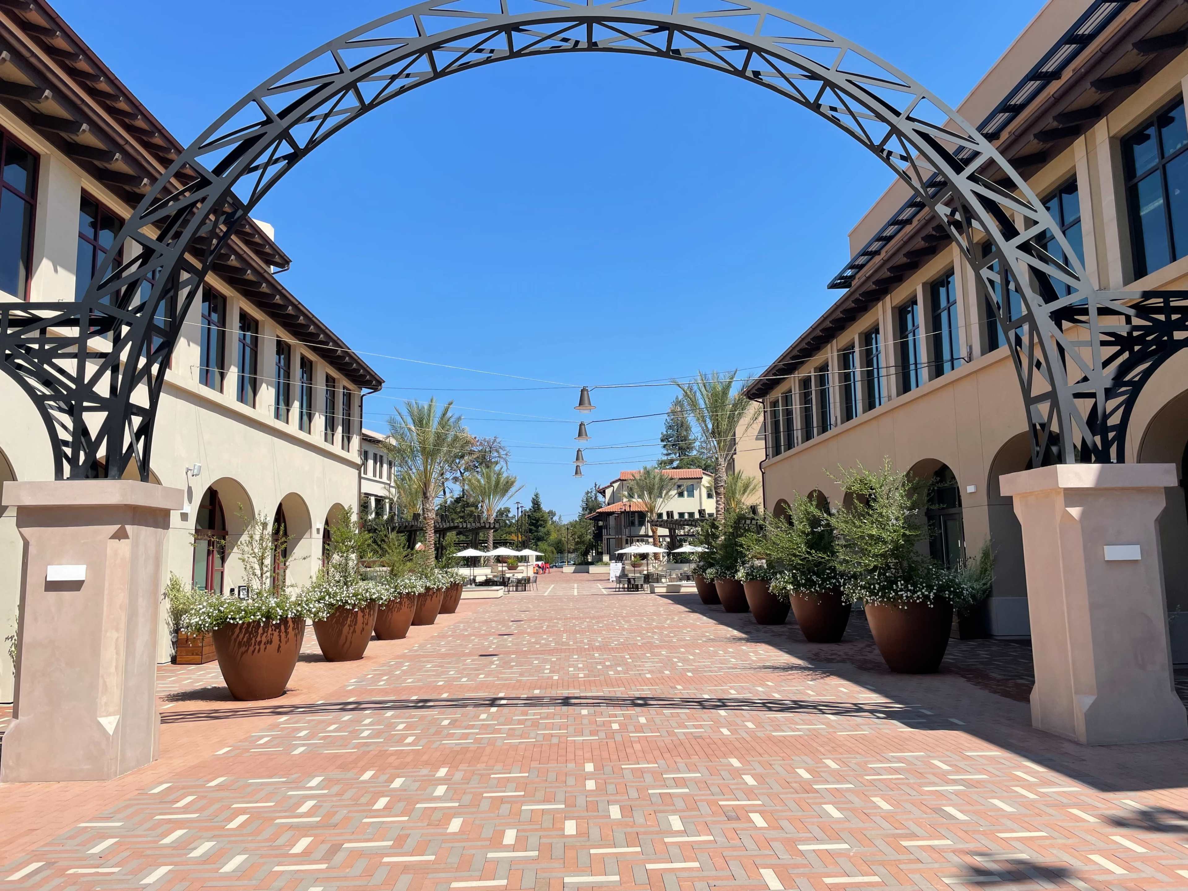 A wide brick pathway framed by a metal arch, flanked by large planters and two buildings on either side under a clear blue sky.