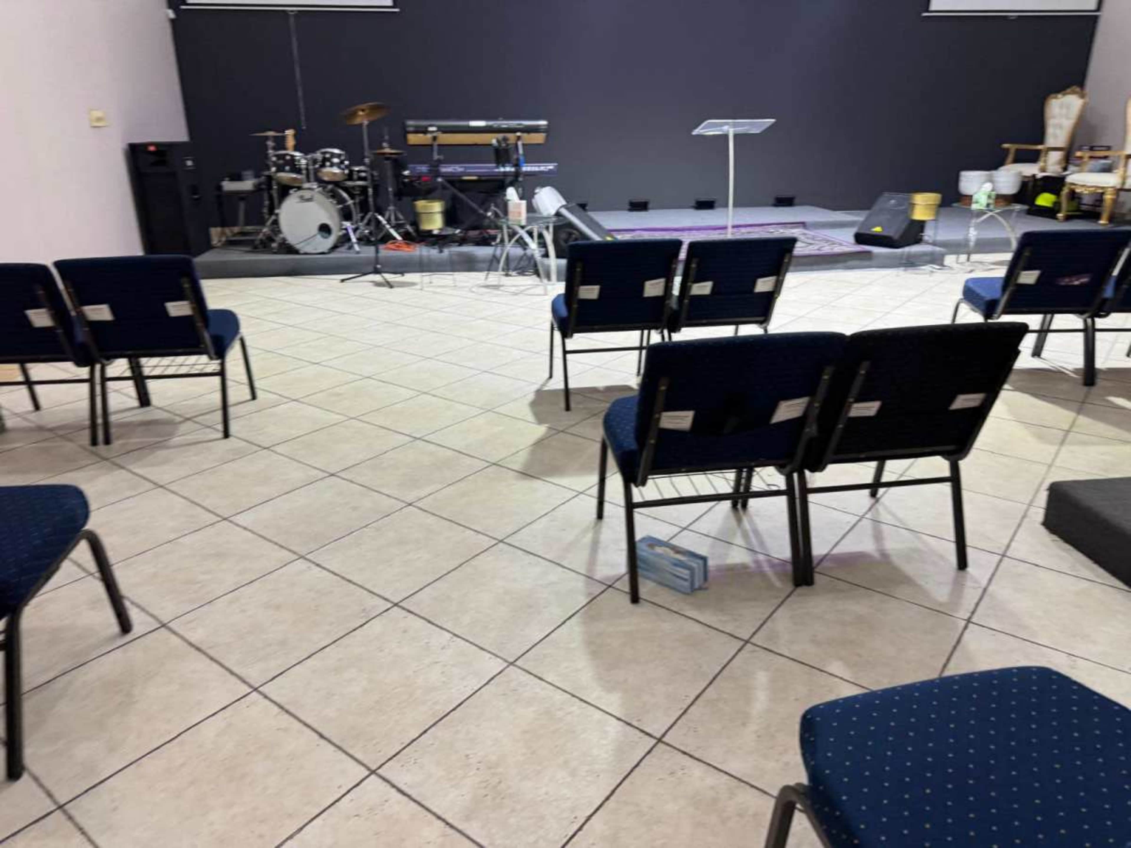 The image shows a spacious, empty room arranged with chairs facing a stage with a music setup, a podium, and a few musical instruments.