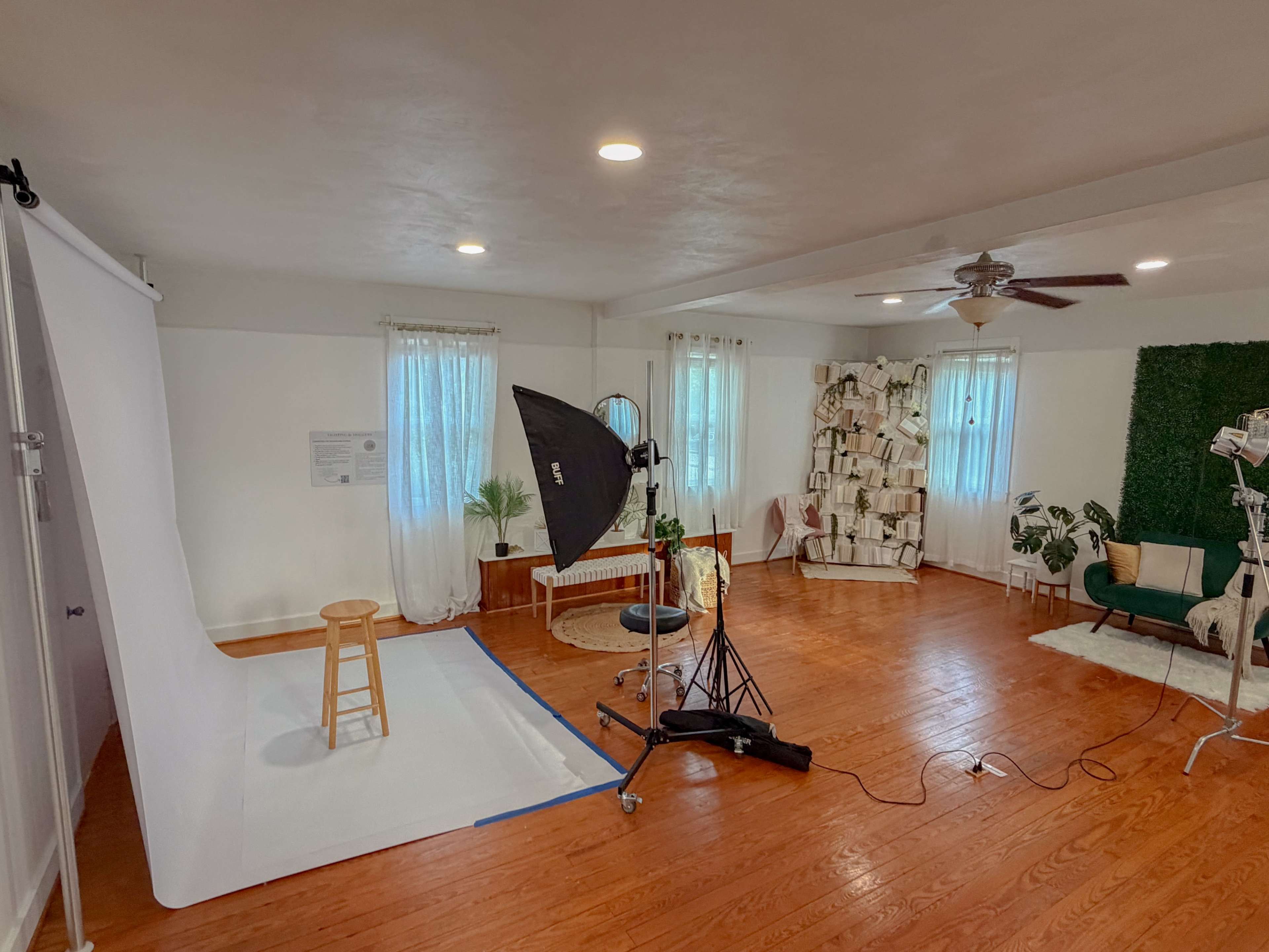 The image shows a photography studio setup with a white backdrop, lighting equipment, a stool, and a green accent wall adorned with books and plants.