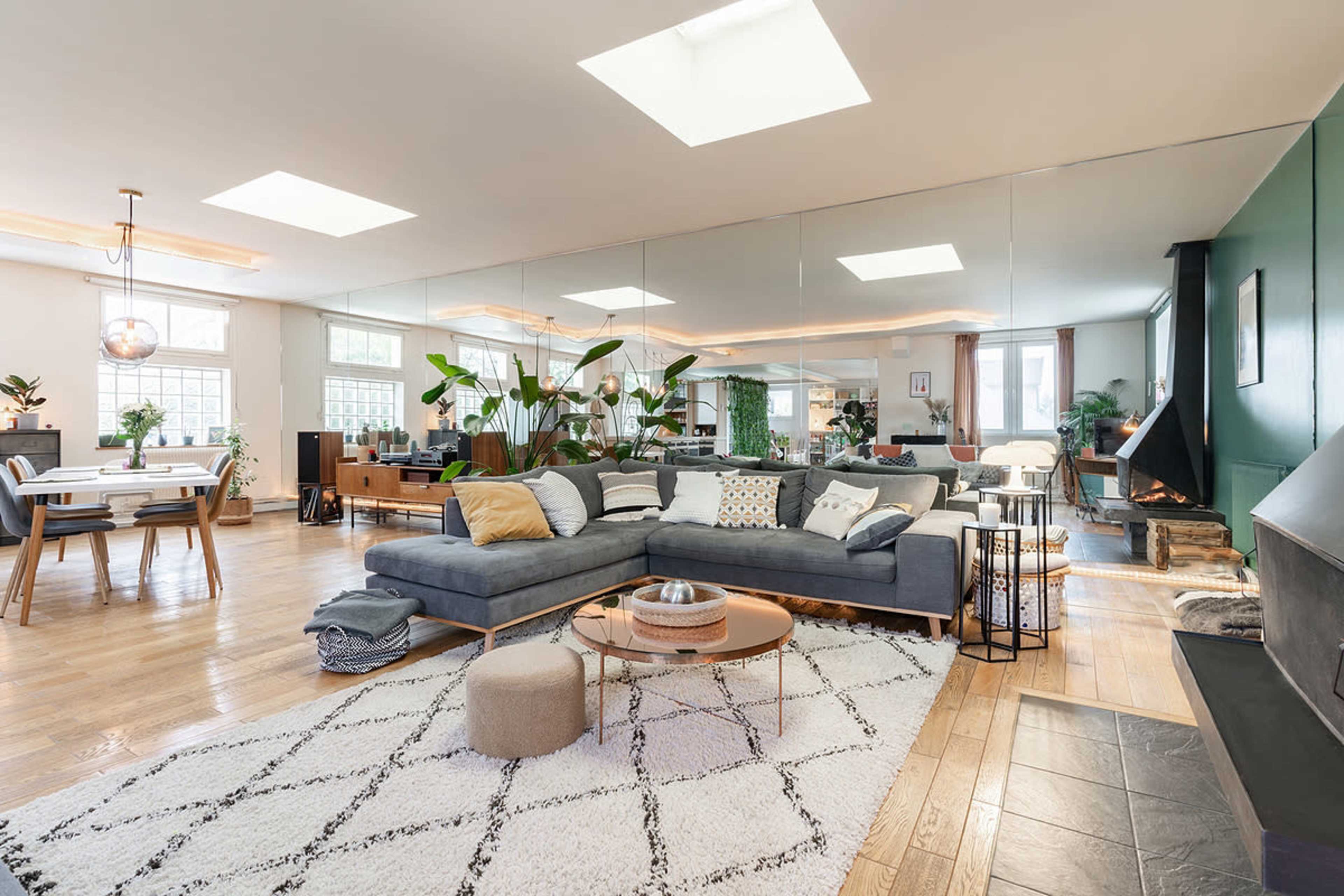 The image shows a spacious living area featuring a sectional sofa, a coffee table, and large windows, with plants and a dining area in the background.