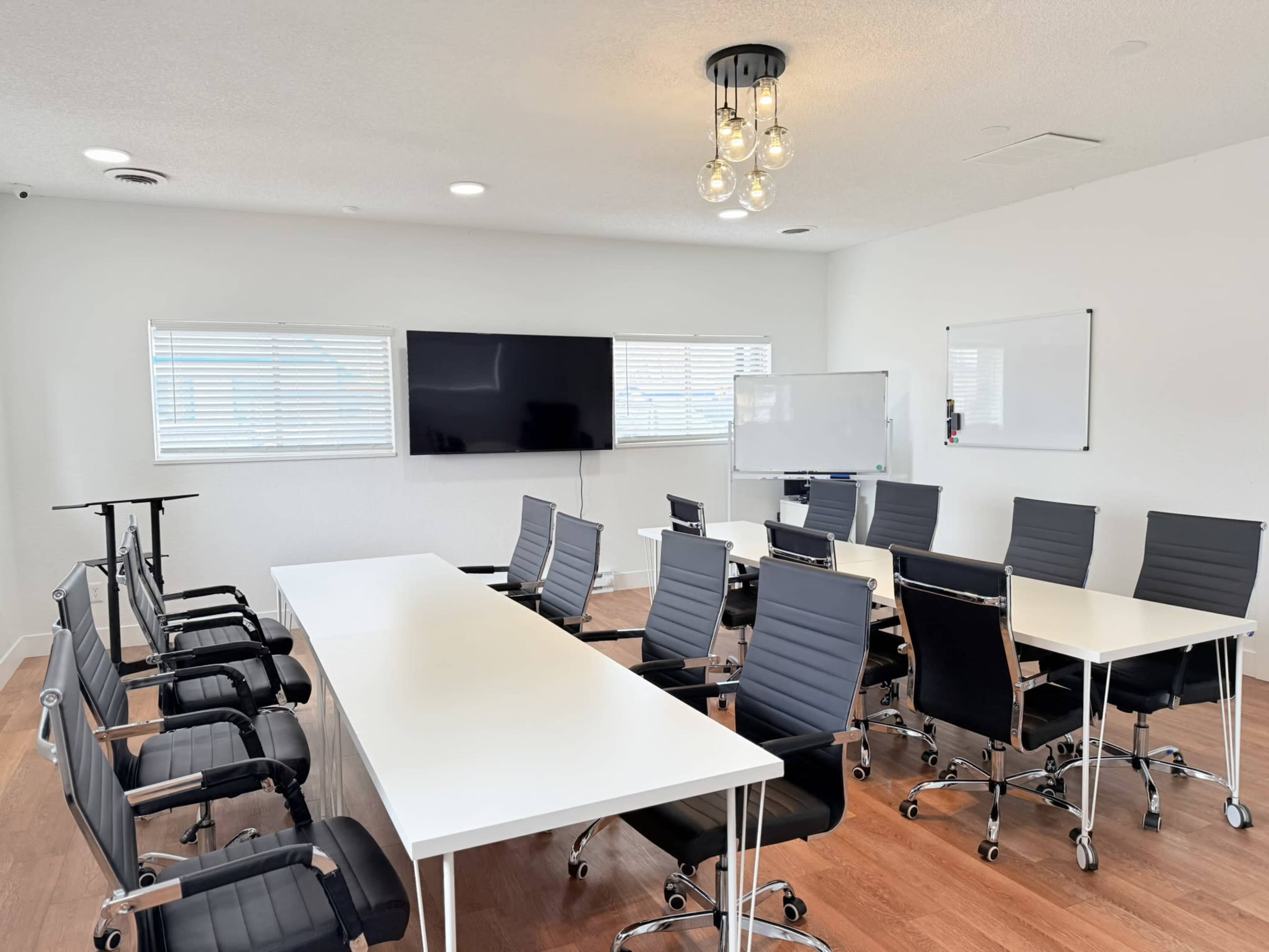 A modern conference room features a long white table surrounded by black office chairs, with two large screens on the walls and a whiteboard.