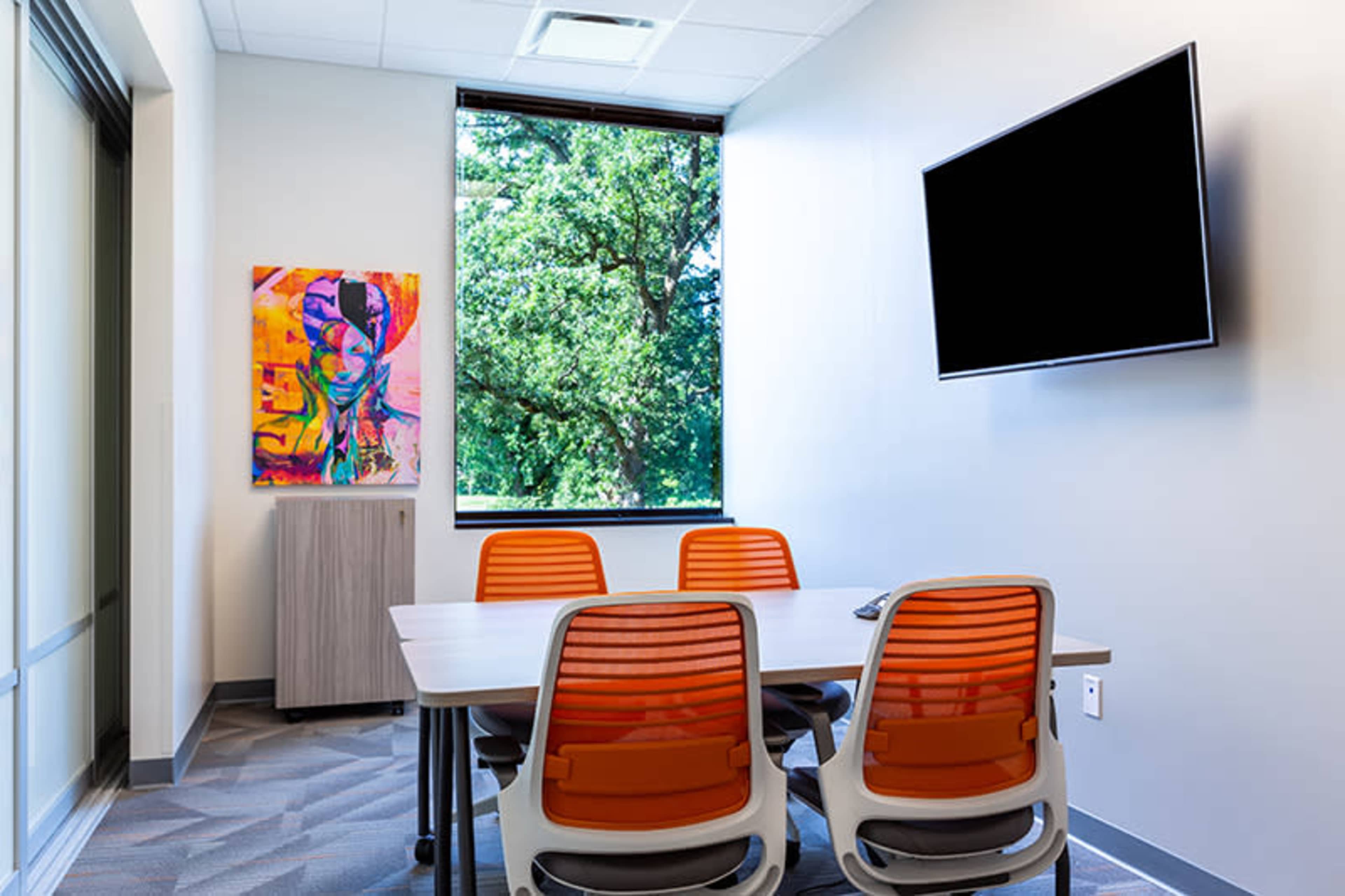 A conference room features a table surrounded by four orange chairs, a wall-mounted television, and a large window overlooking greenery outside.