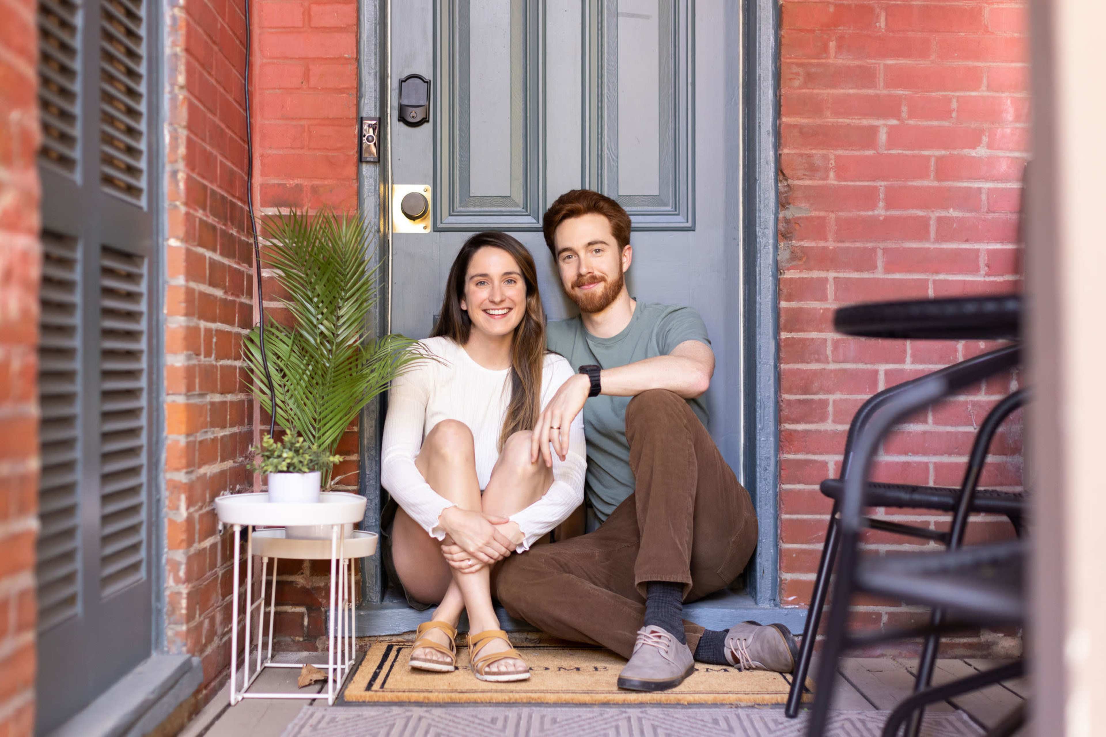 A couple sits on the front porch beside a potted plant, smiling at the camera in front of a brick wall and a gray door.