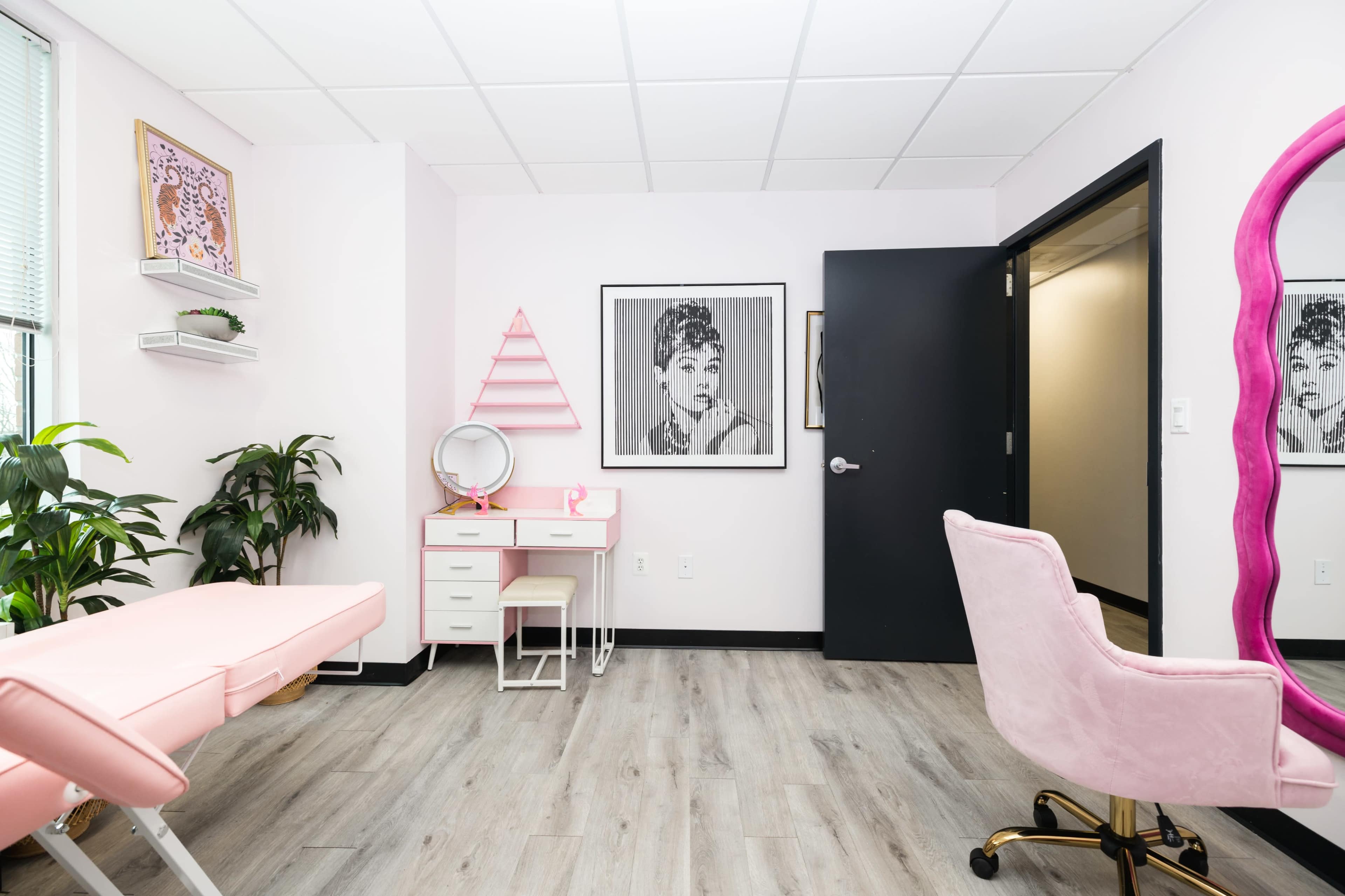 A brightly lit room with pink and white decor, featuring a makeup station, a pink chair, and artwork on the walls.