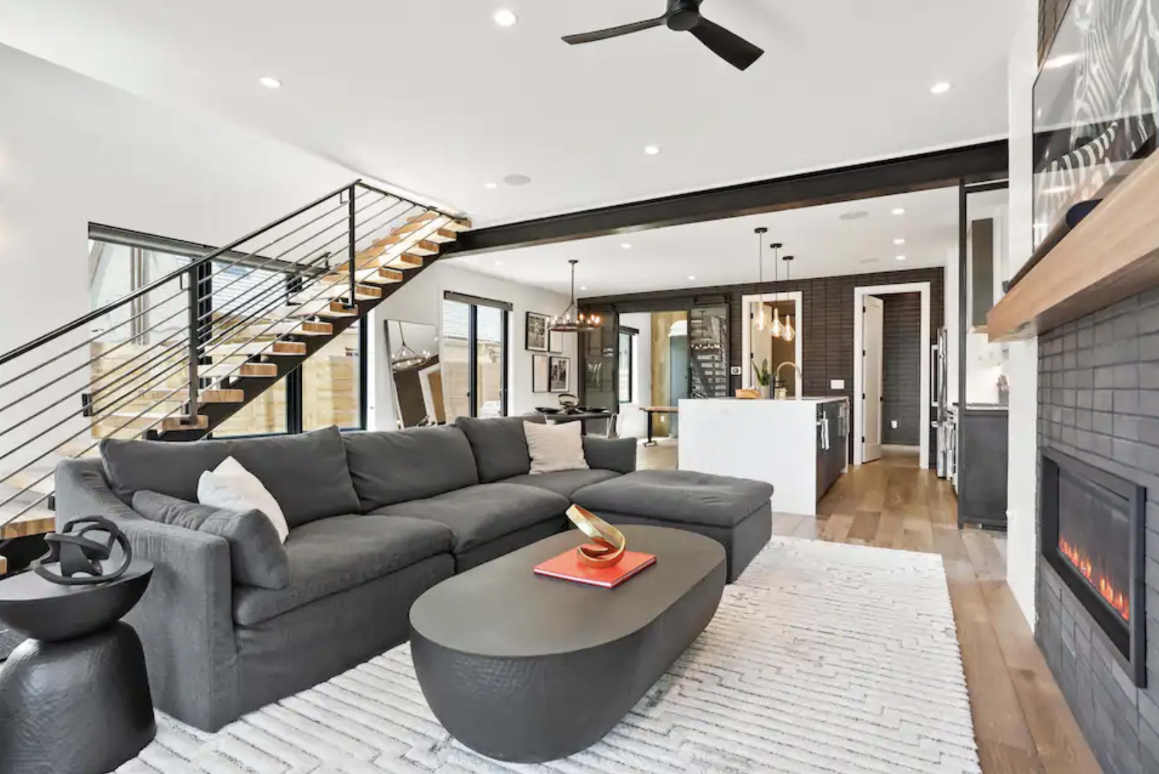 A modern living room features a gray sectional sofa, a round coffee table, a sleek fireplace, and a view of an open kitchen with a staircase leading to the upper level.