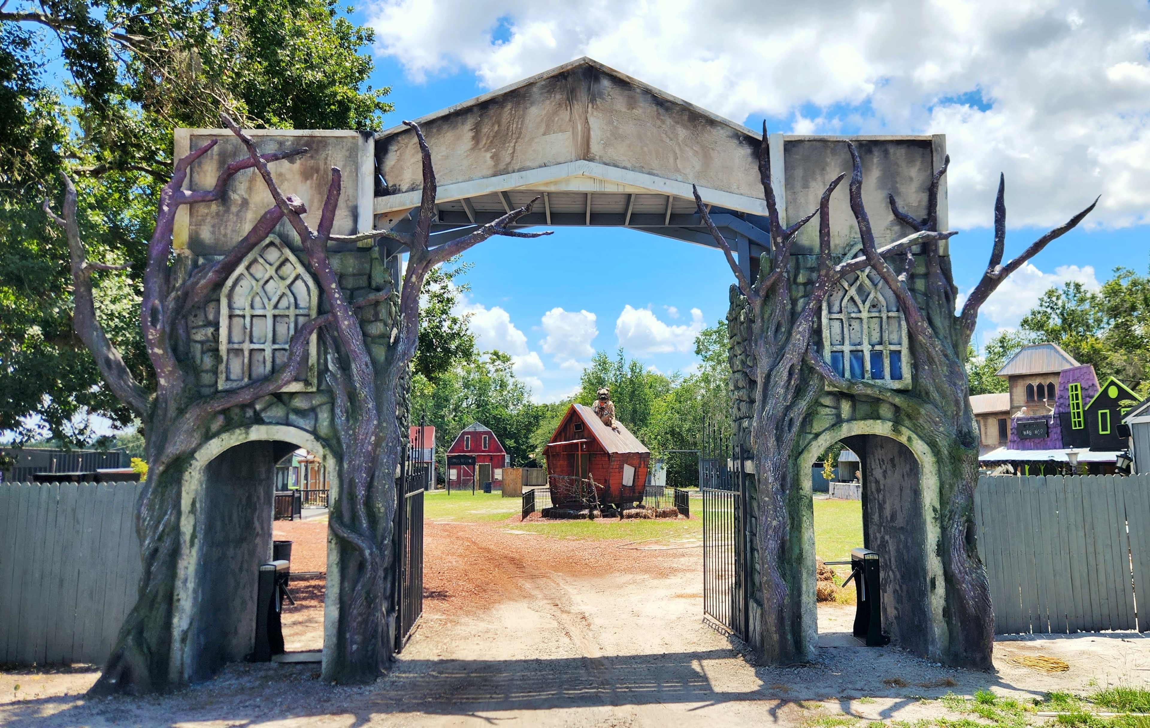 A large archway with tree-like structures at its base, leading into an open area with a few buildings in the background under a partly cloudy sky.