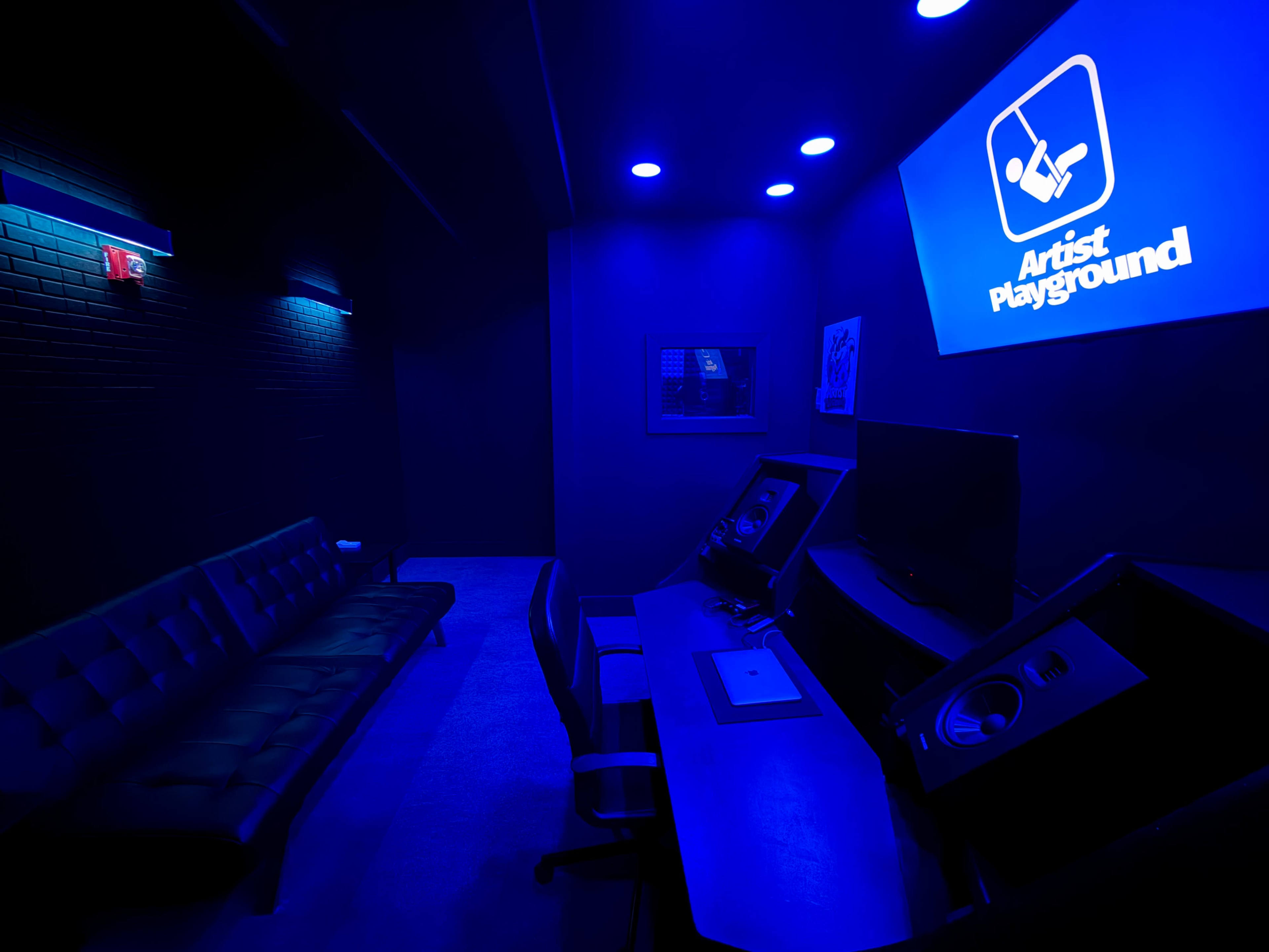 The image shows a dimly lit room with blue lighting, featuring a sleek black sofa, a desk with sound equipment, and a wall-mounted screen displaying the logo "Artist Playground."