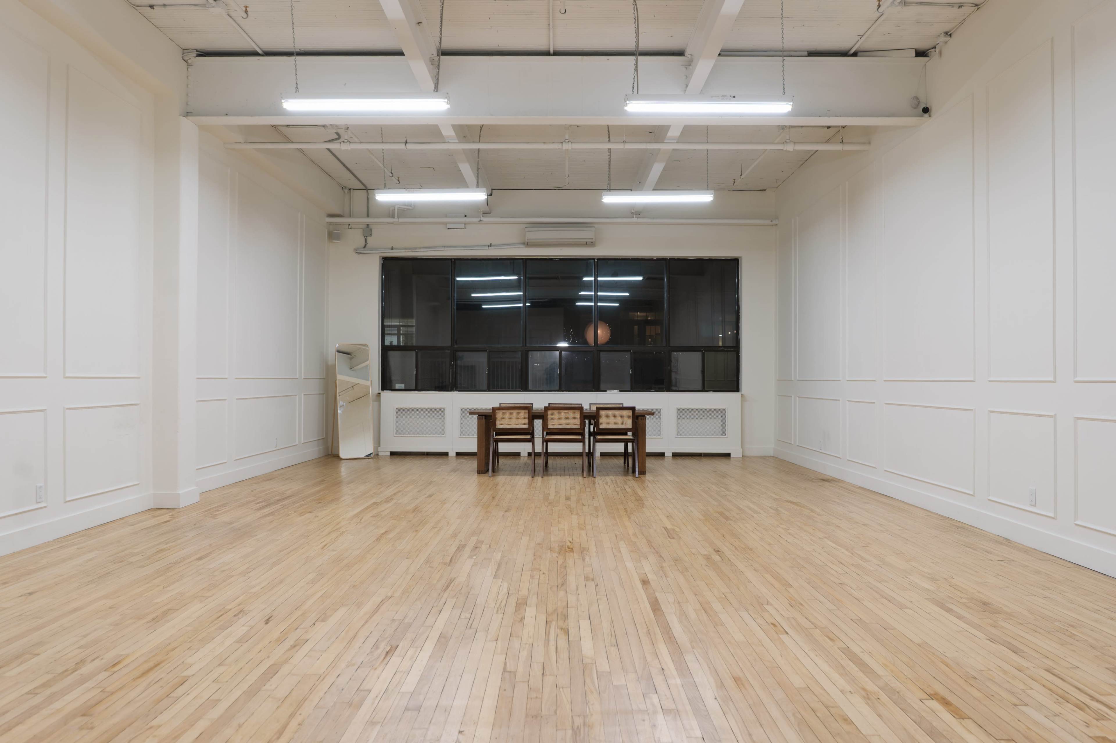 A spacious, empty room featuring hardwood floors, white walls, and a table with chairs positioned in front of a large window.