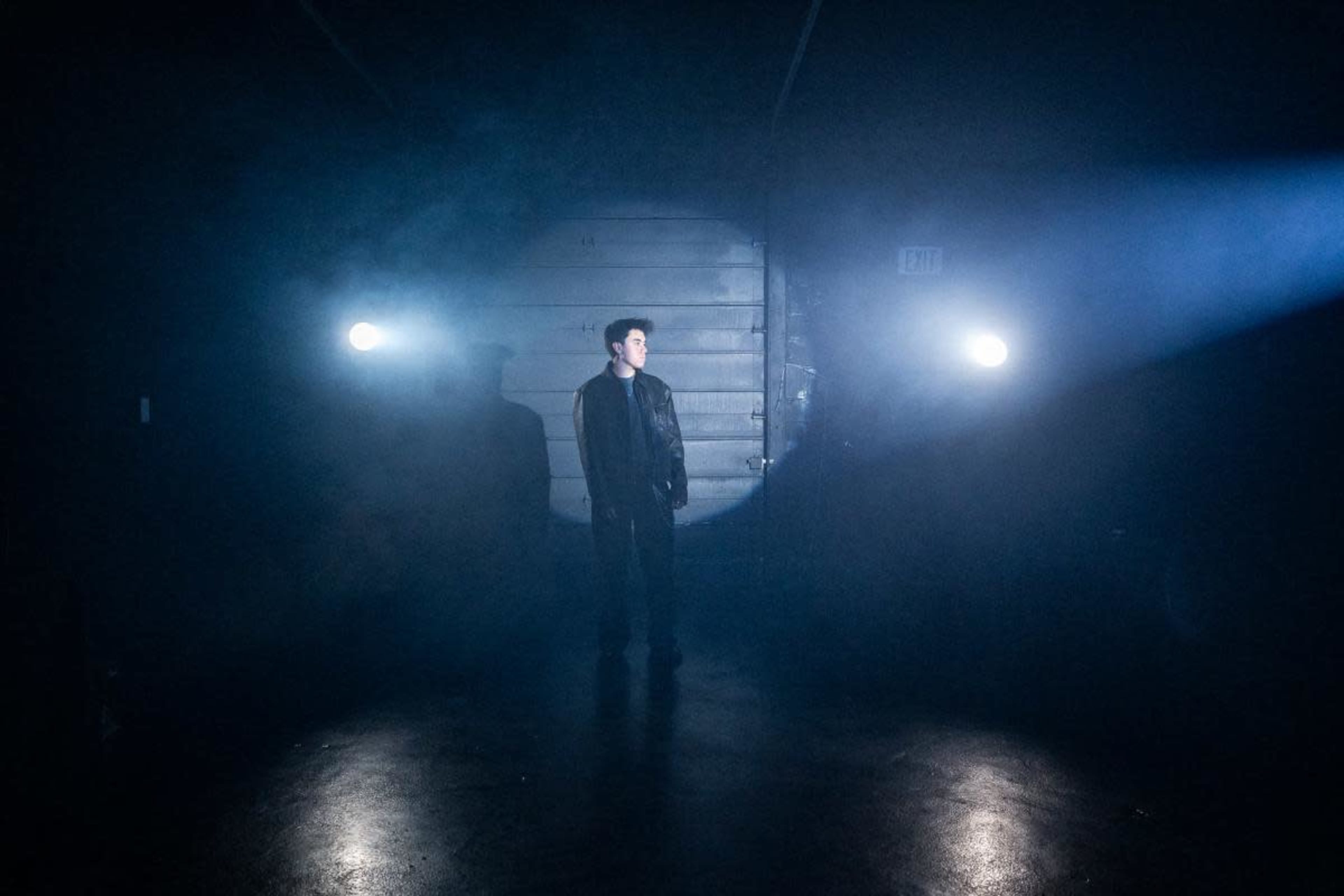 A person stands alone in a dimly lit space with beams of light illuminating them against a shadowy background.