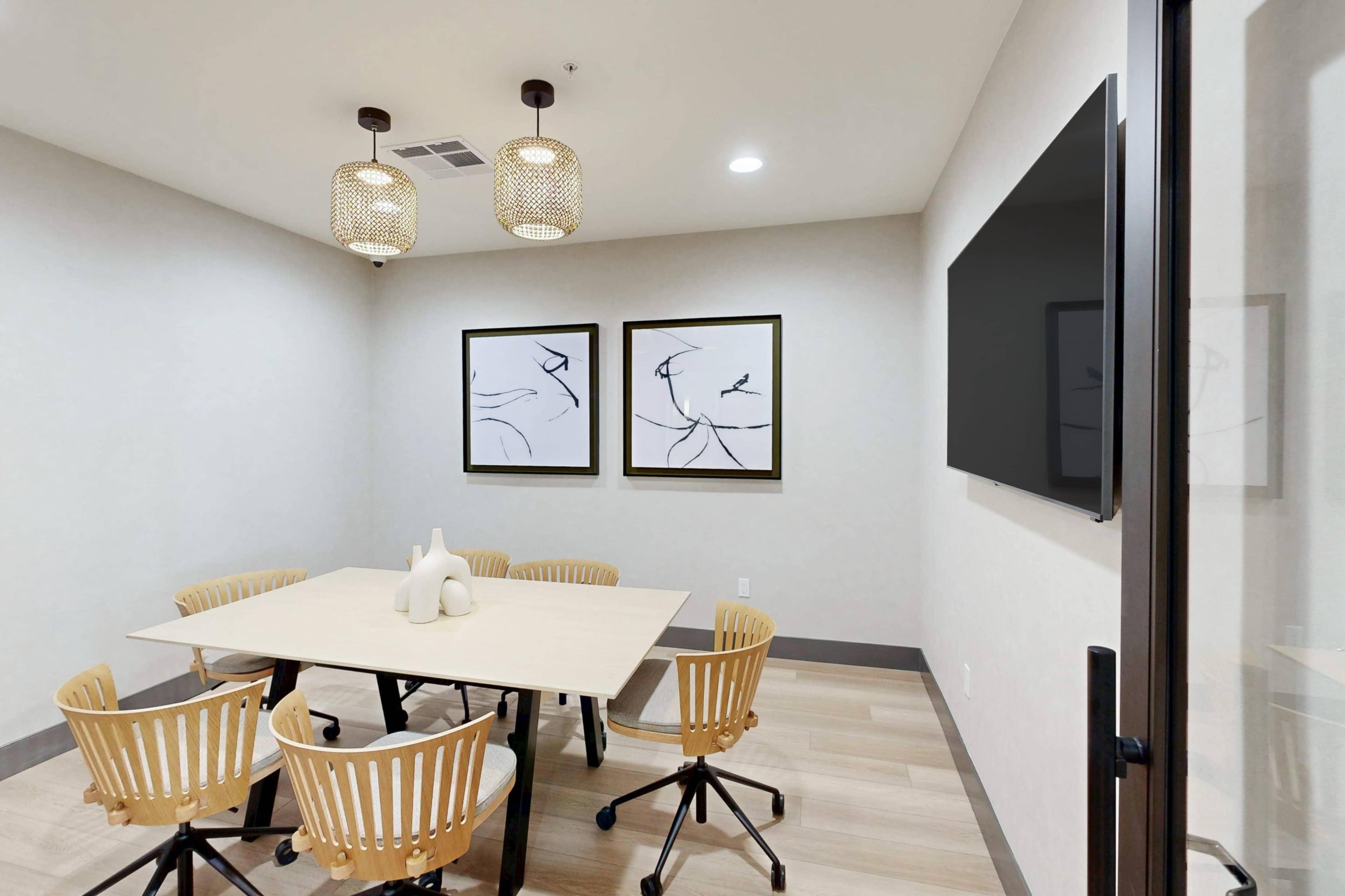 The image shows a modern meeting room with a large table surrounded by six rolling chairs, two framed artworks on the wall, and a television mounted on the opposite wall.