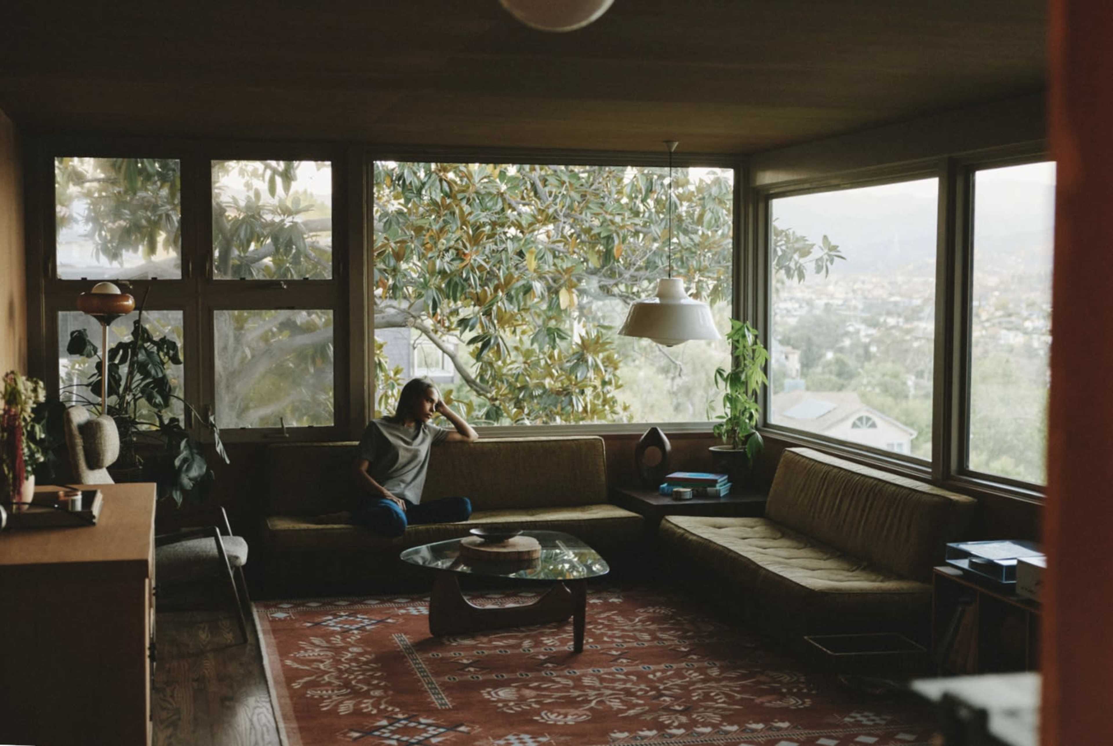 A person sits thoughtfully on a couch in a spacious living room with large windows overlooking a view of greenery and distant hills.