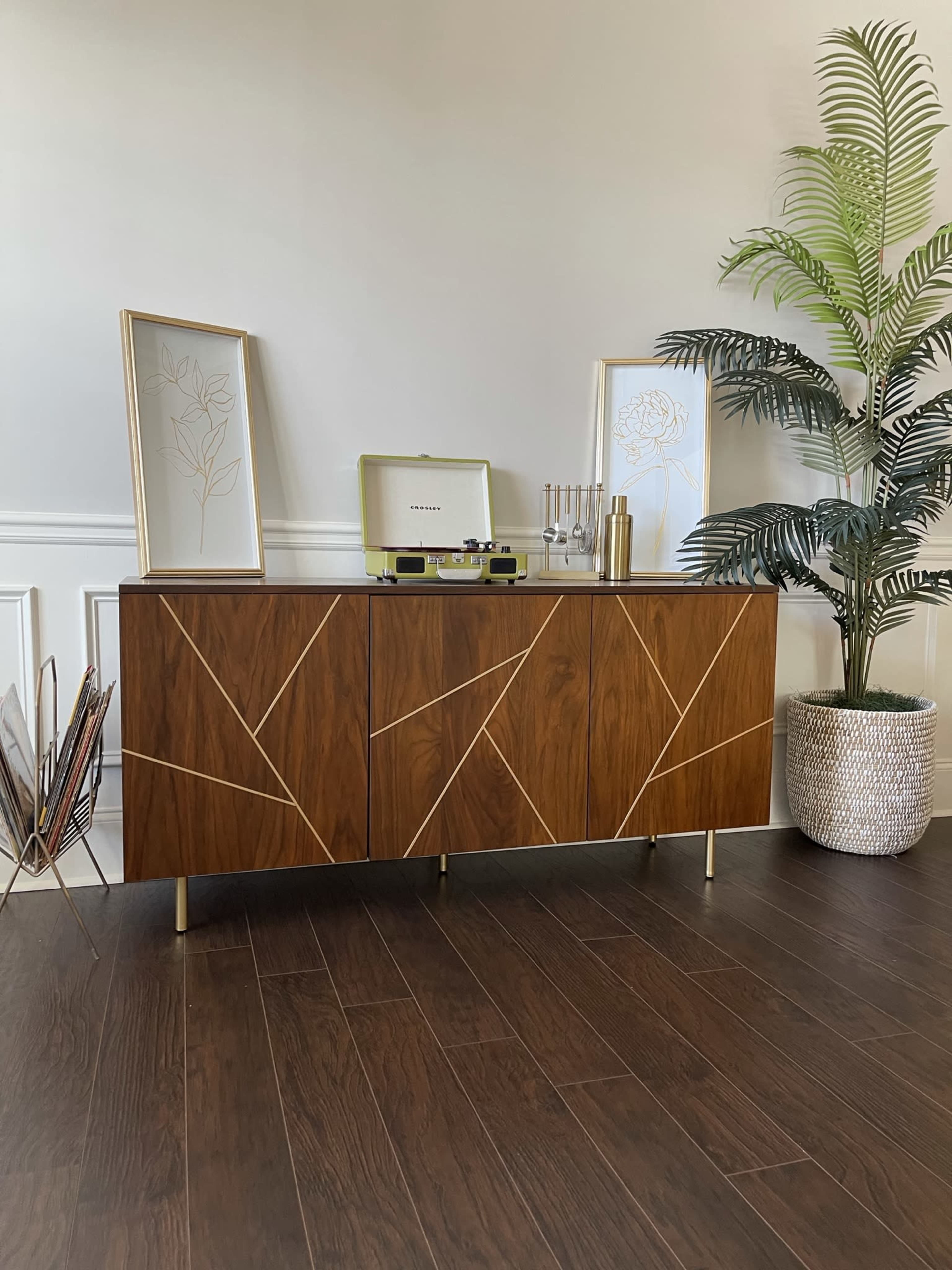 A wooden sideboard with decorative lines sits against a wall, accompanied by a record player and framed botanical art, while a potted plant stands beside it.