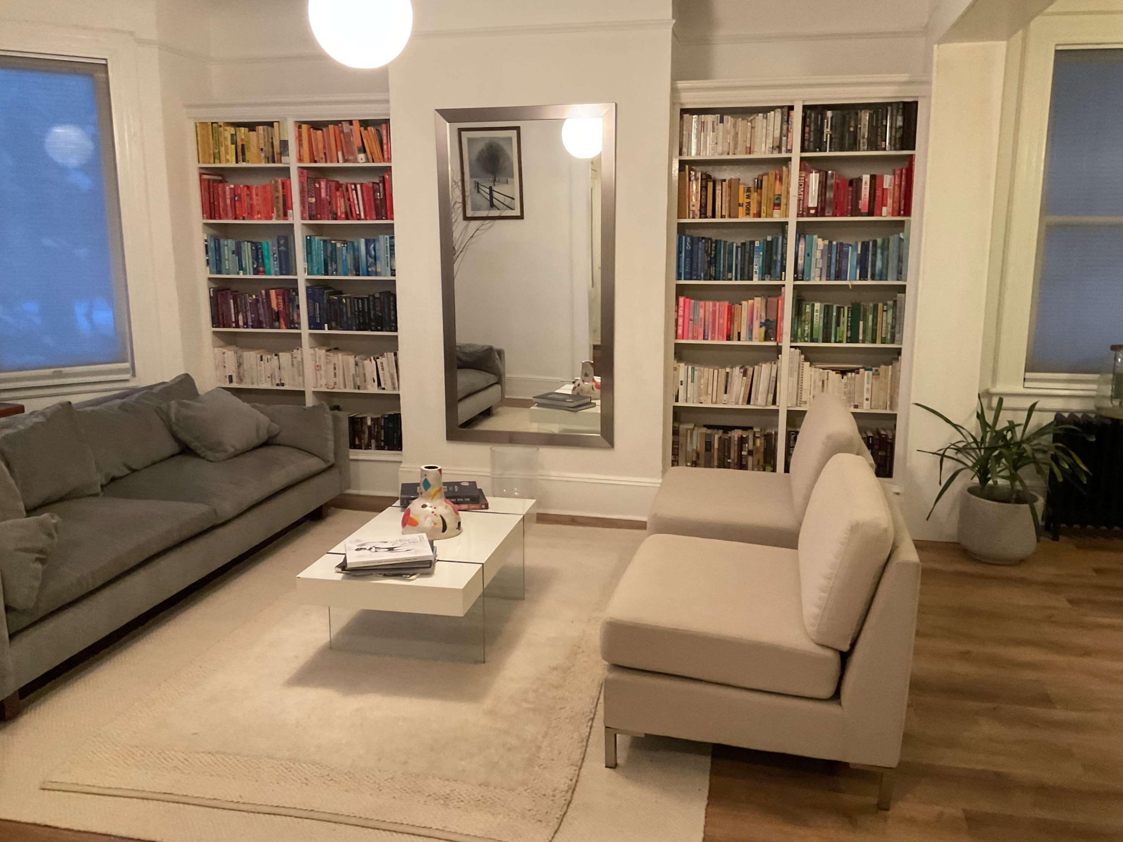 A modern living room features a gray sofa and a pair of light-colored chairs facing a white coffee table, with bookshelves organized by color and a large mirror on the wall.