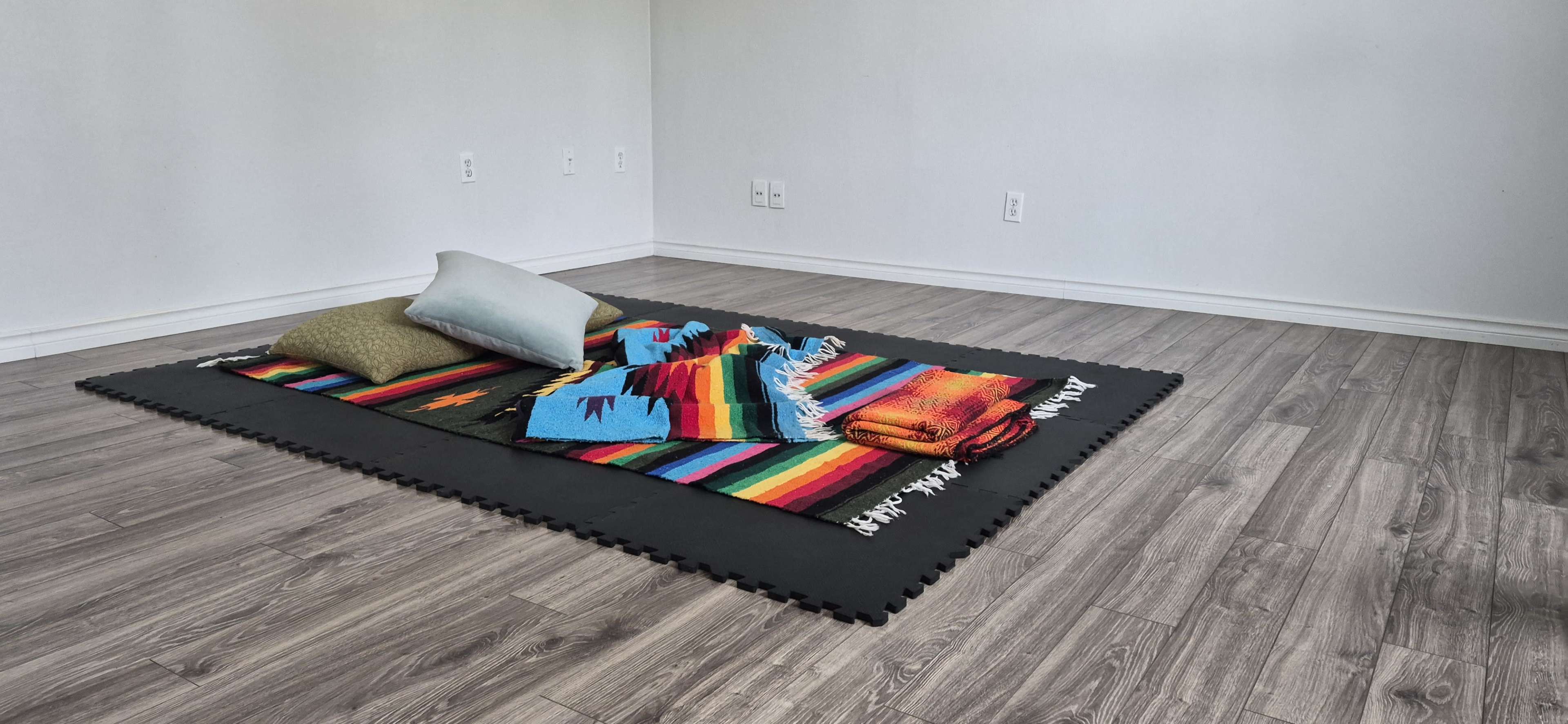 A colorful woven blanket and pillows are arranged on a black mat in an empty room with wooden flooring.