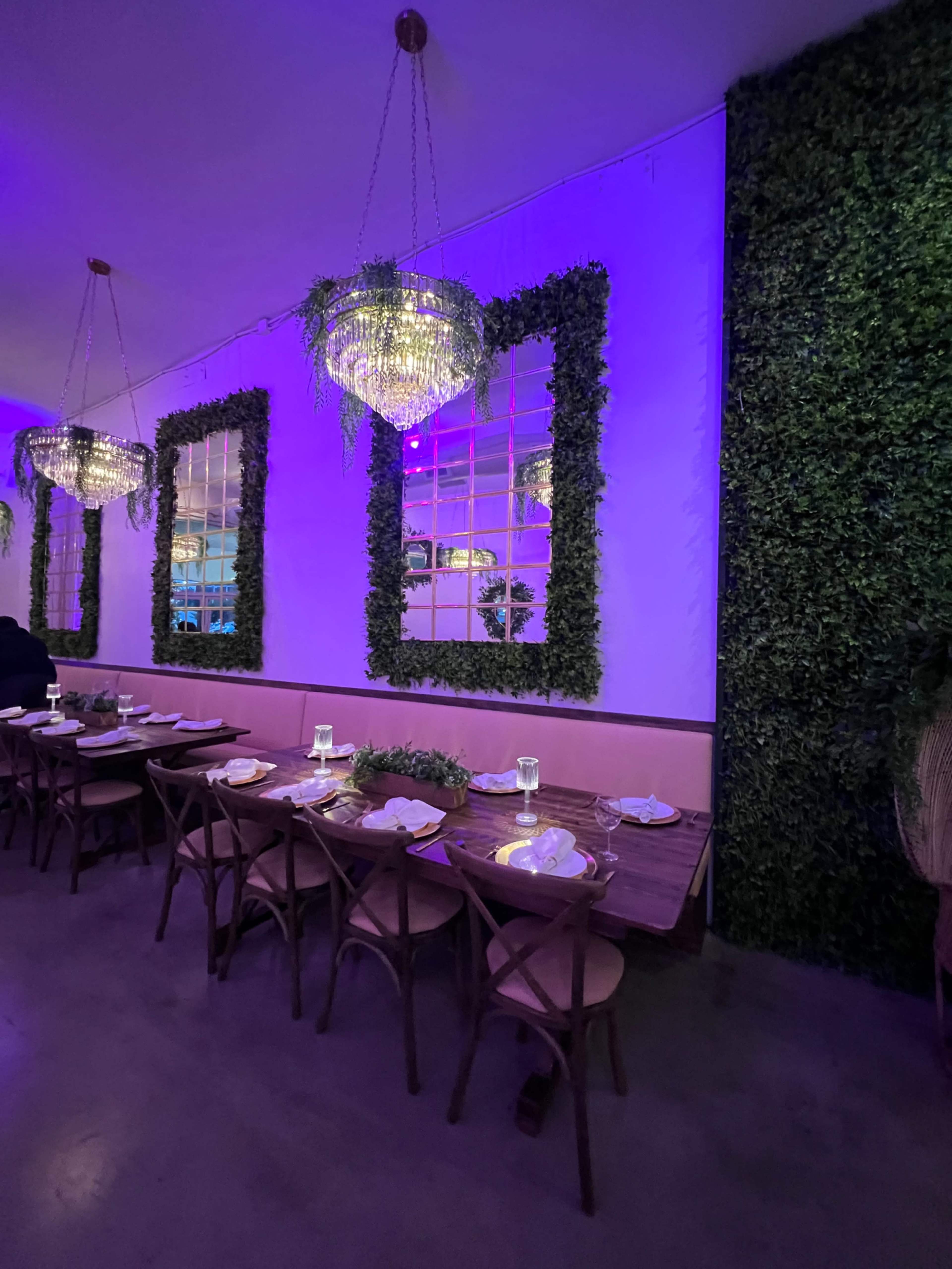 The image shows a restaurant interior featuring a long wooden table set for dining, with purple lighting, chandeliers, and a wall adorned with greenery.