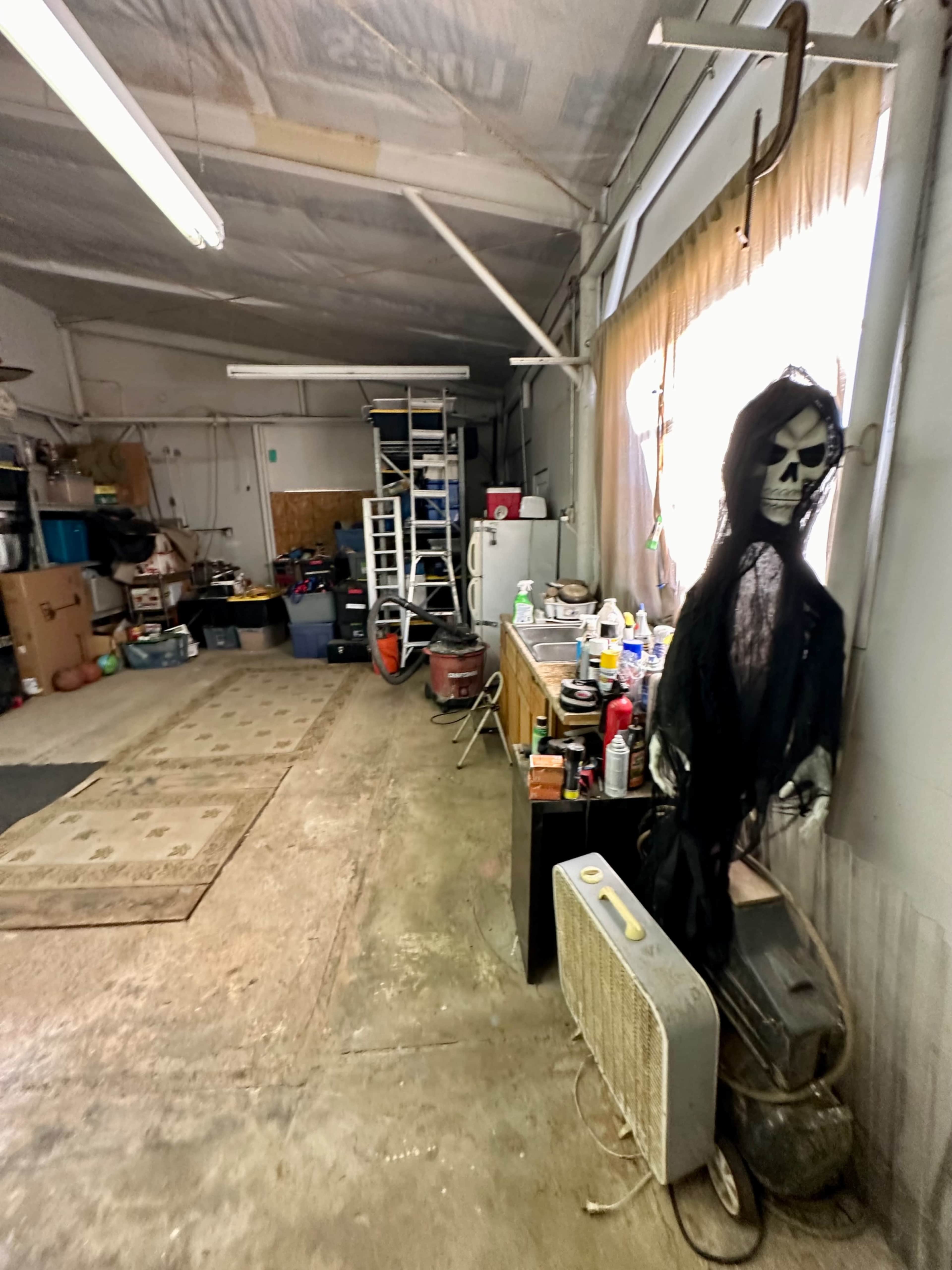 The image shows a cluttered garage or storage area with various items, including a shelf ladder, boxes, cleaning supplies, and a ghostly figure dressed in black near a fan.
