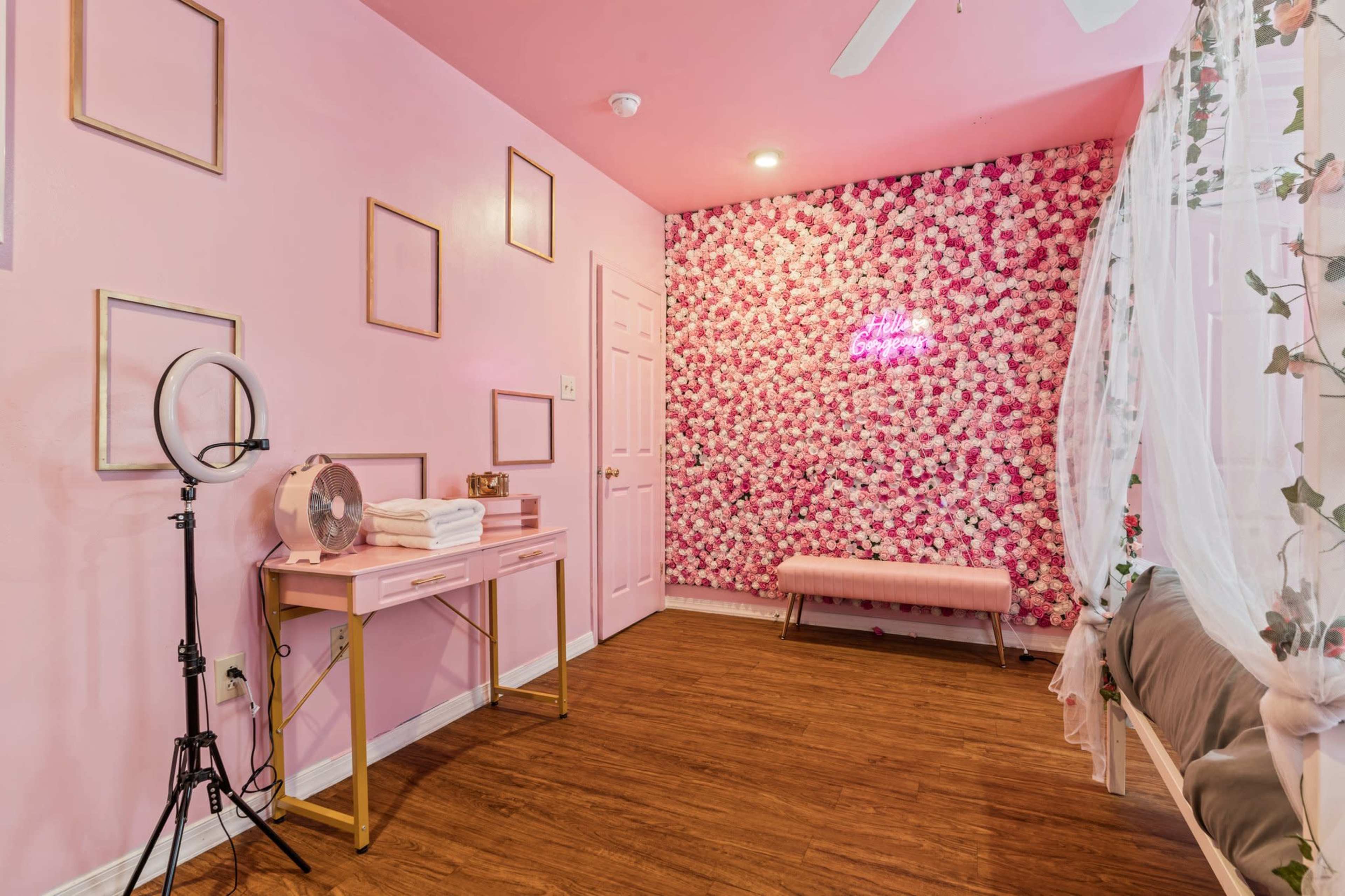 The room features pink walls adorned with floral decor, a seating bench, a mirrored table, and a lighted sign, creating a bright, well-decorated space.