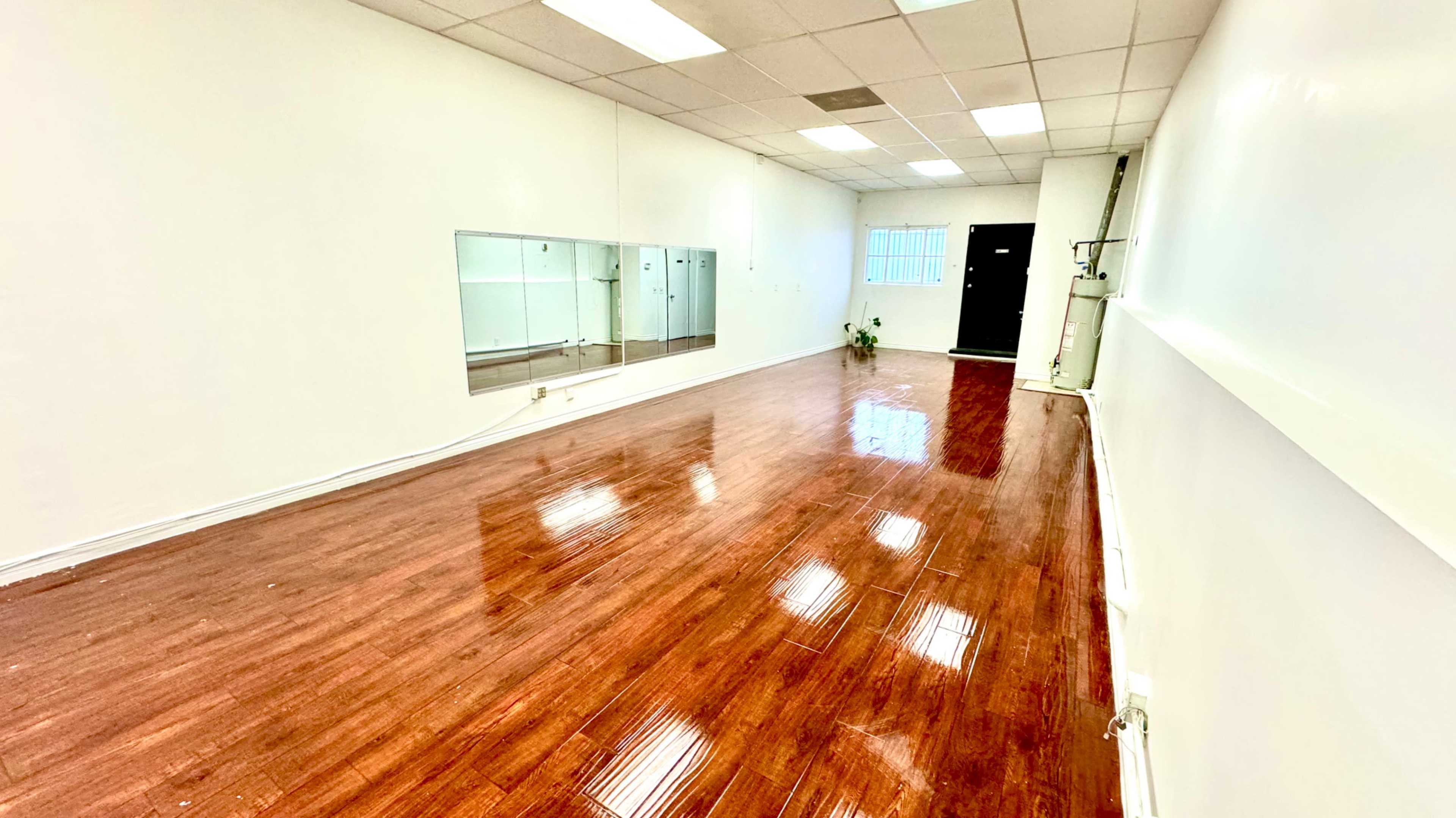 A spacious, well-lit room with polished wooden flooring, mirrors on one wall, and an entrance at the far end.