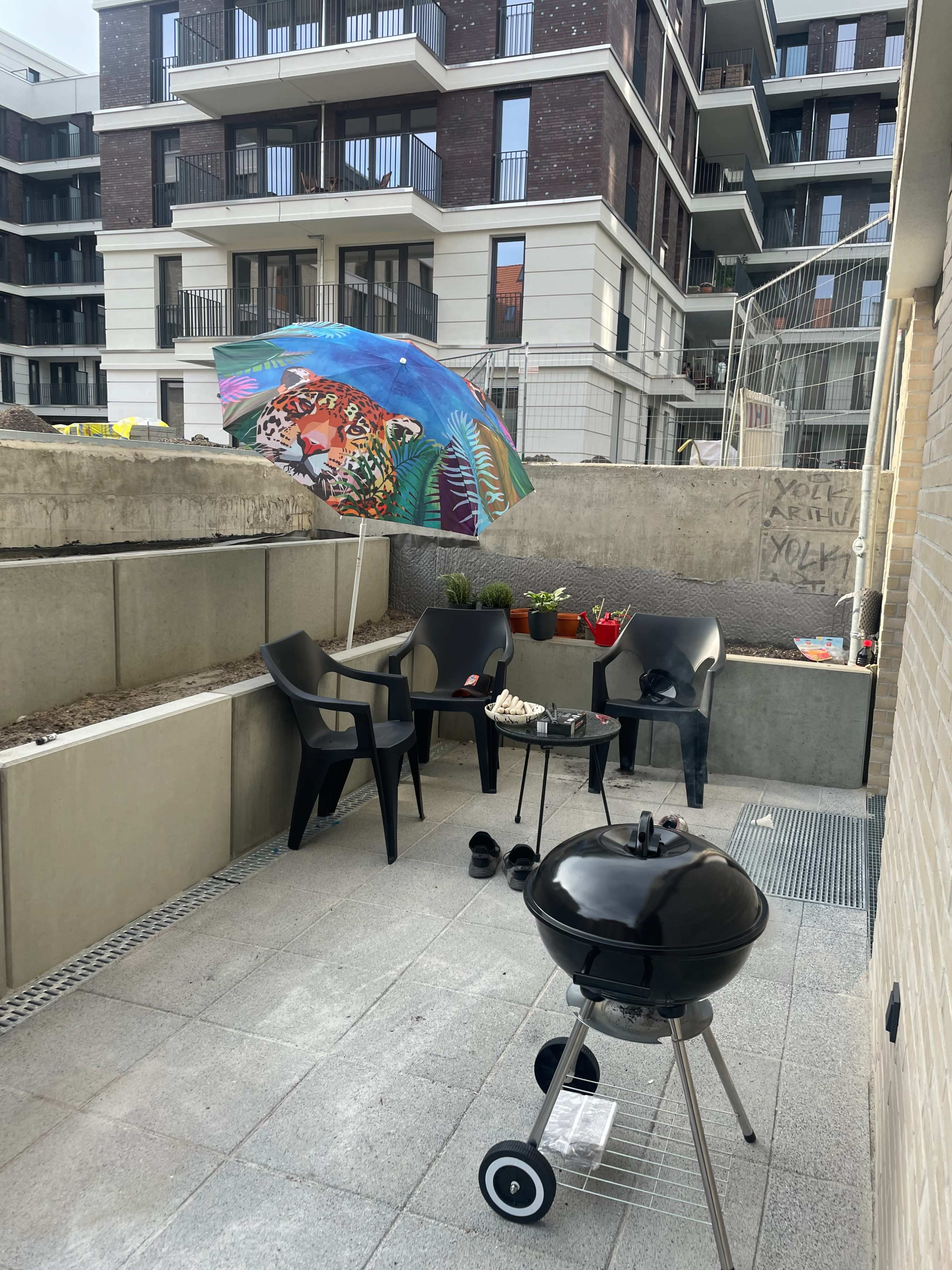 A small balcony features a black charcoal grill, a colorful umbrella, chairs, and potted plants surrounded by modern apartment buildings.