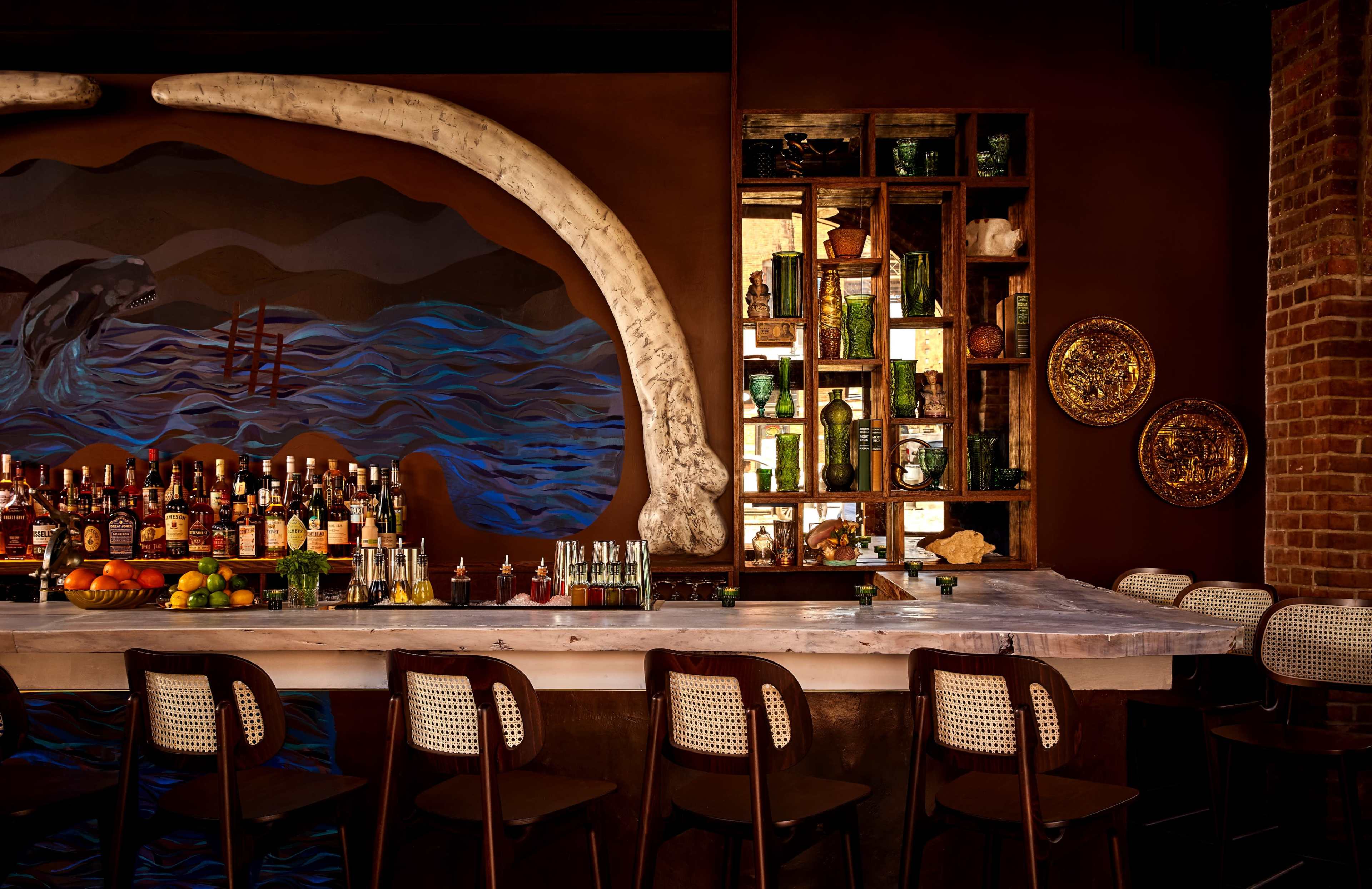 The image shows a bar with a marble countertop, a display of various bottles behind it, and a large whale bone mounted on the wall next to a colorful mural of water and ships.