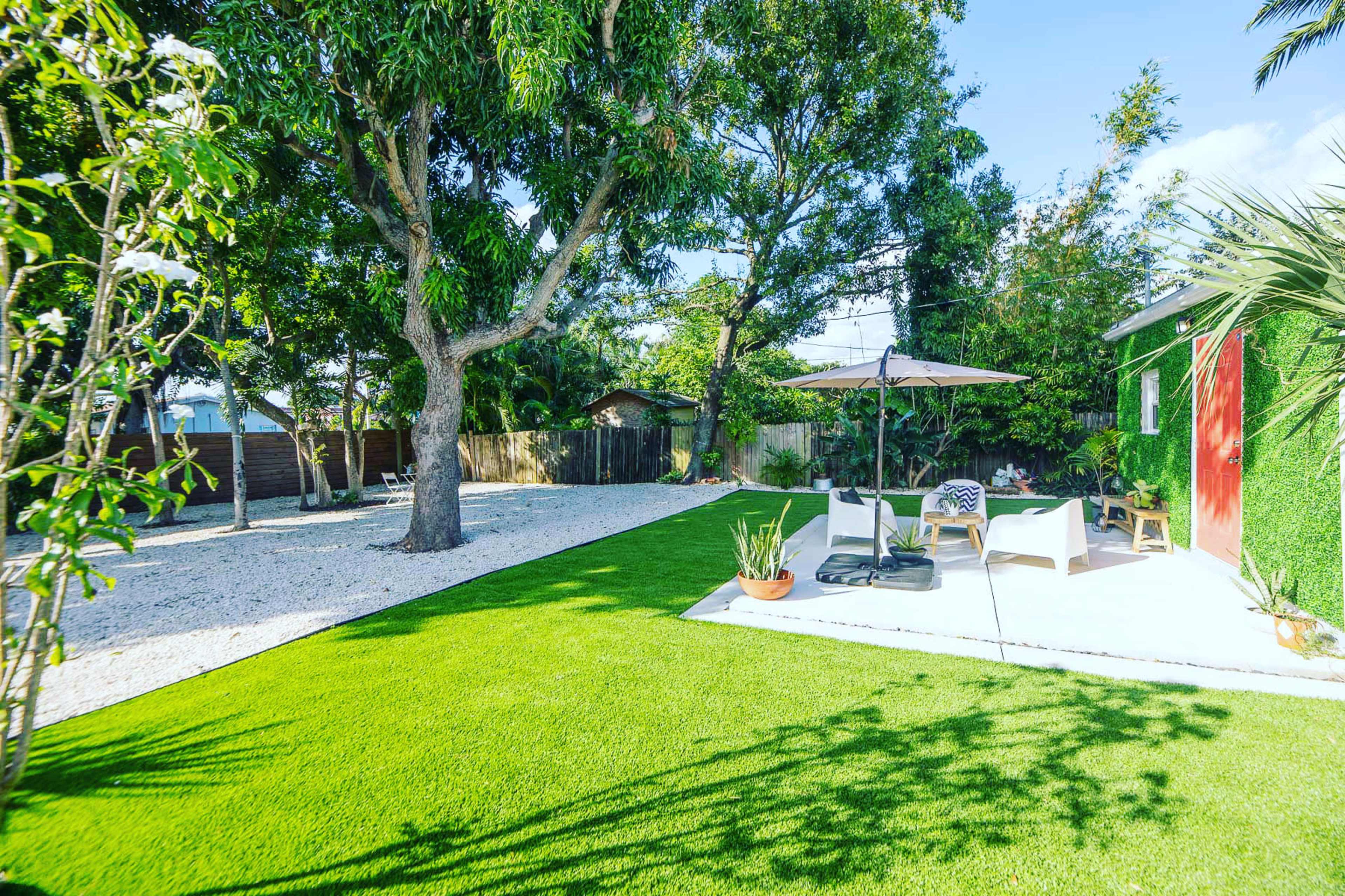 The image shows a well-maintained backyard featuring a grassy area, a patio with seating, and trees surrounded by decorative plants.