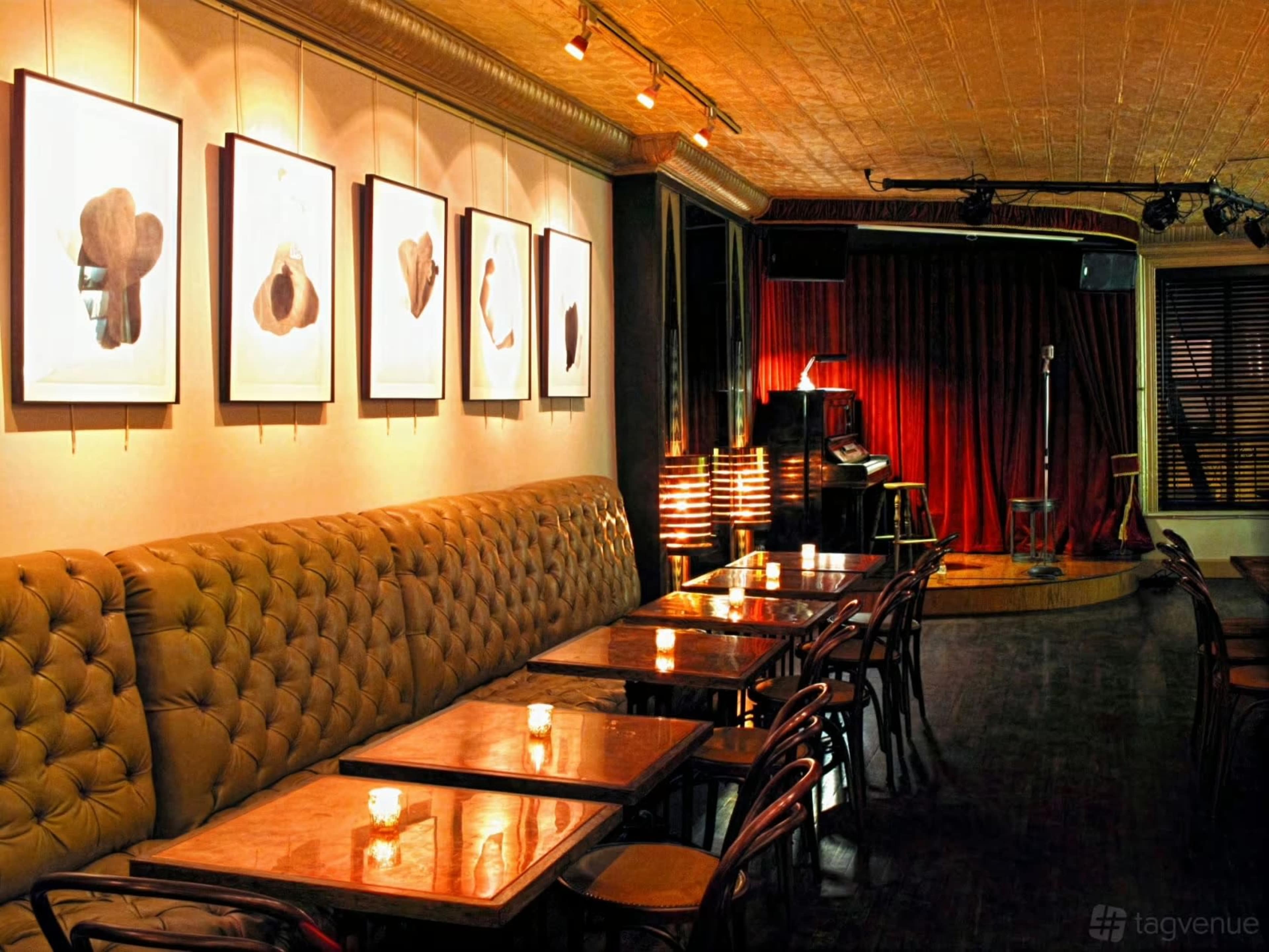 The image shows a cozy performance venue with a stage, seating area featuring wooden tables and chairs, and framed artwork on the walls.