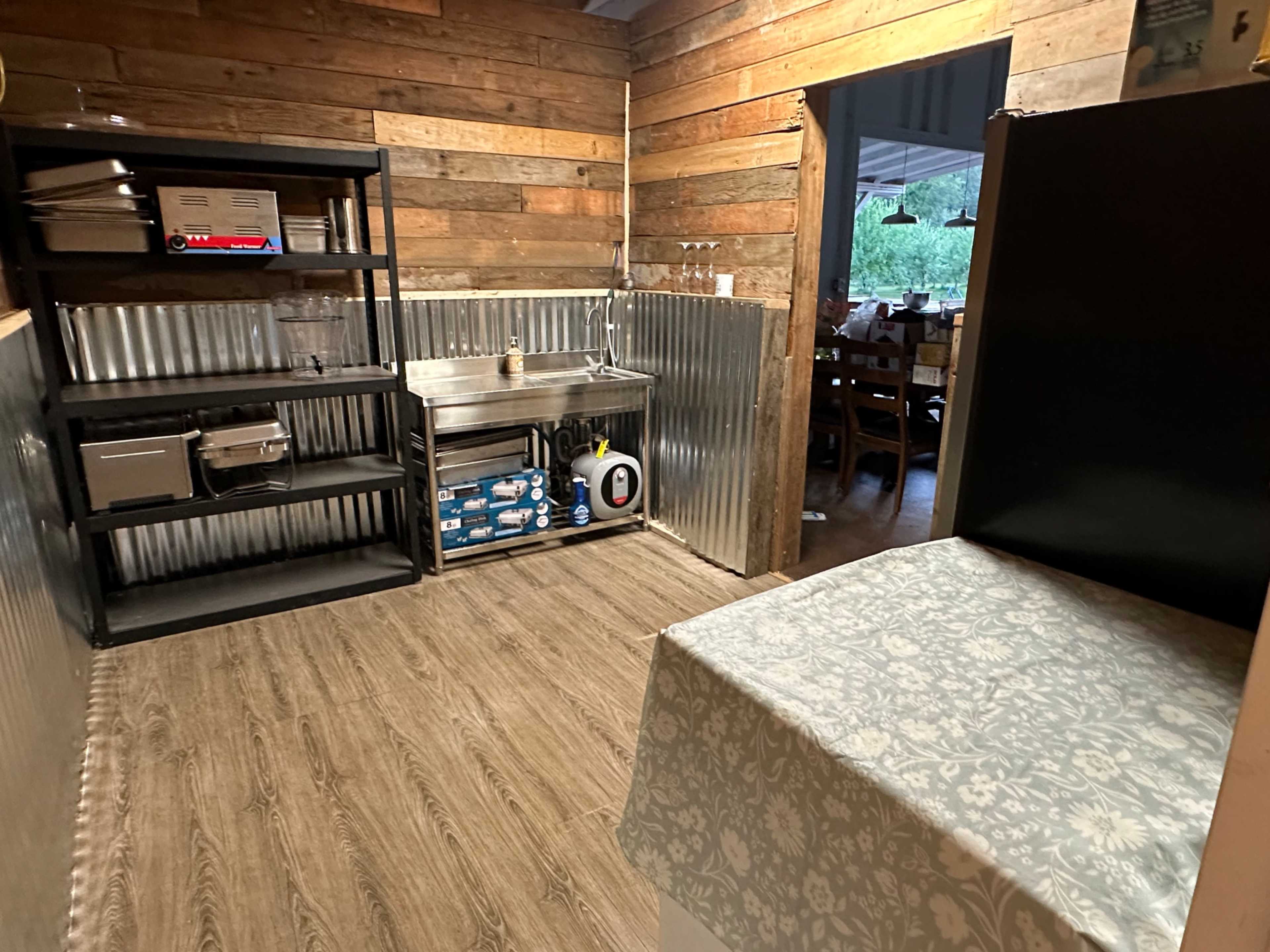 A rustic kitchen with wooden walls, a stainless steel work table, and shelves containing kitchen equipment.