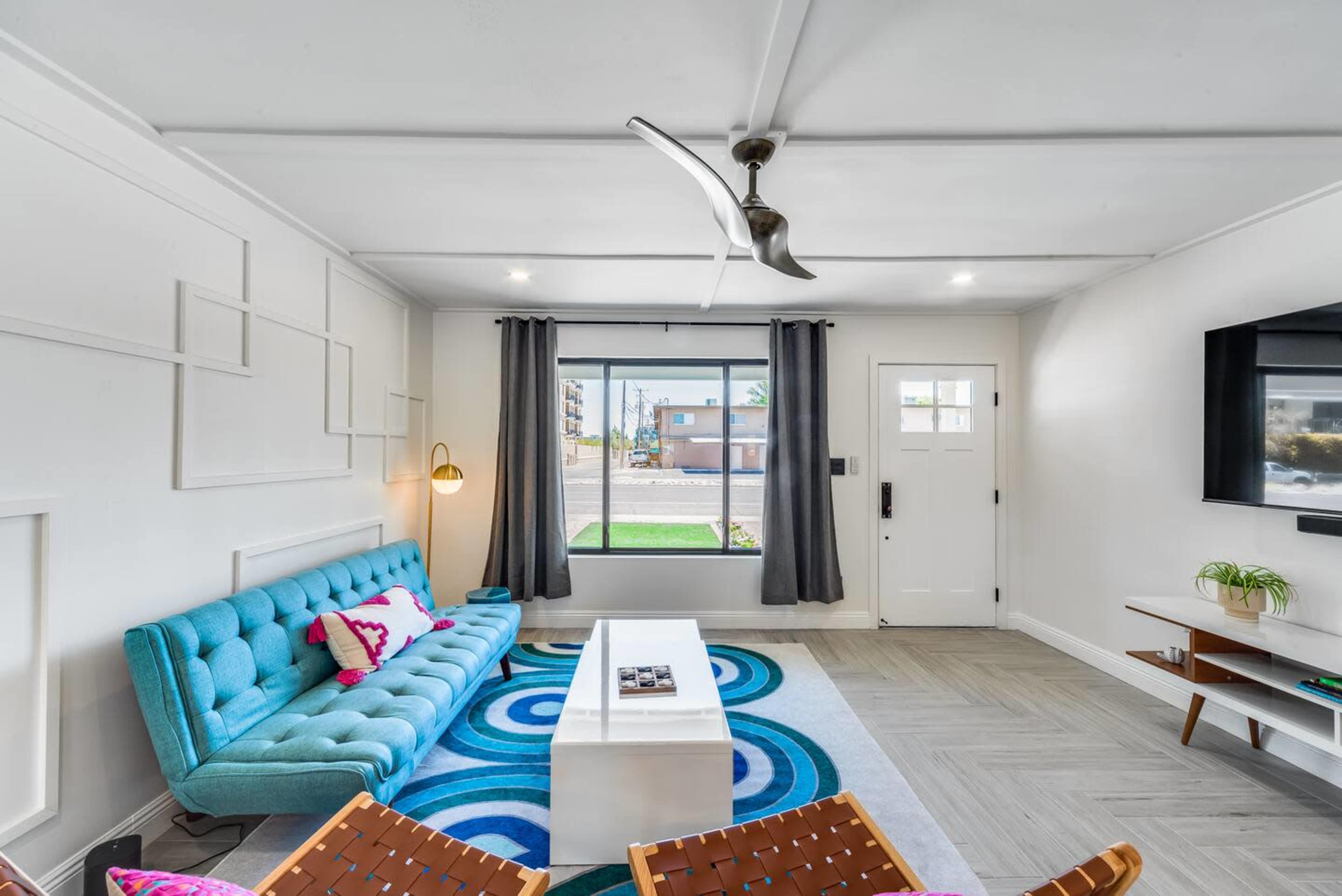 The living room features a blue sofa, a circular patterned rug, and a large window with gray curtains, along with a coffee table and wall-mounted television.