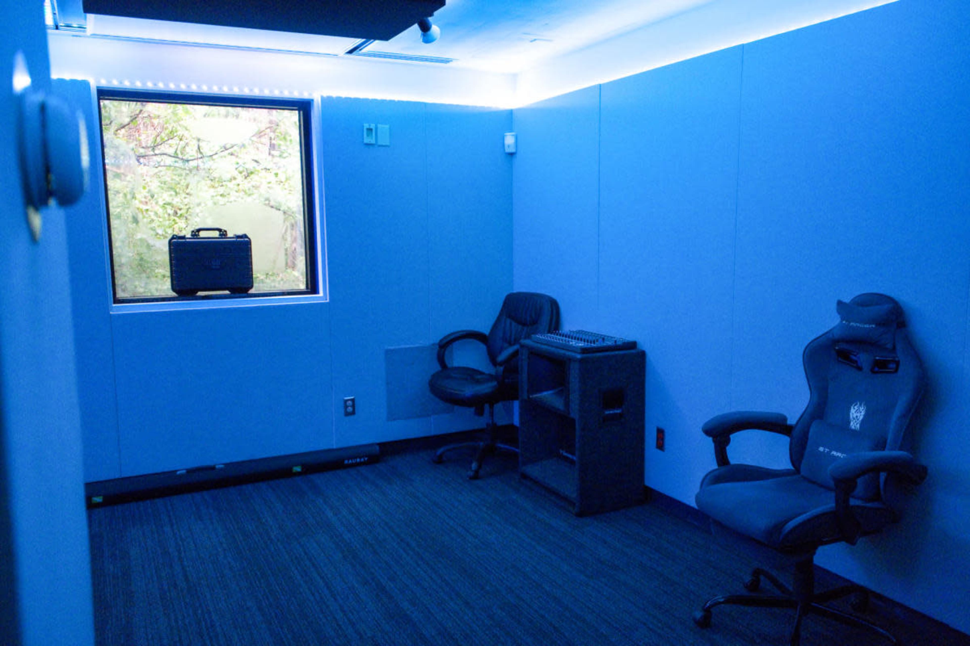 The image shows a small, blue-walled room with two chairs, a window, and equipment on a desk.