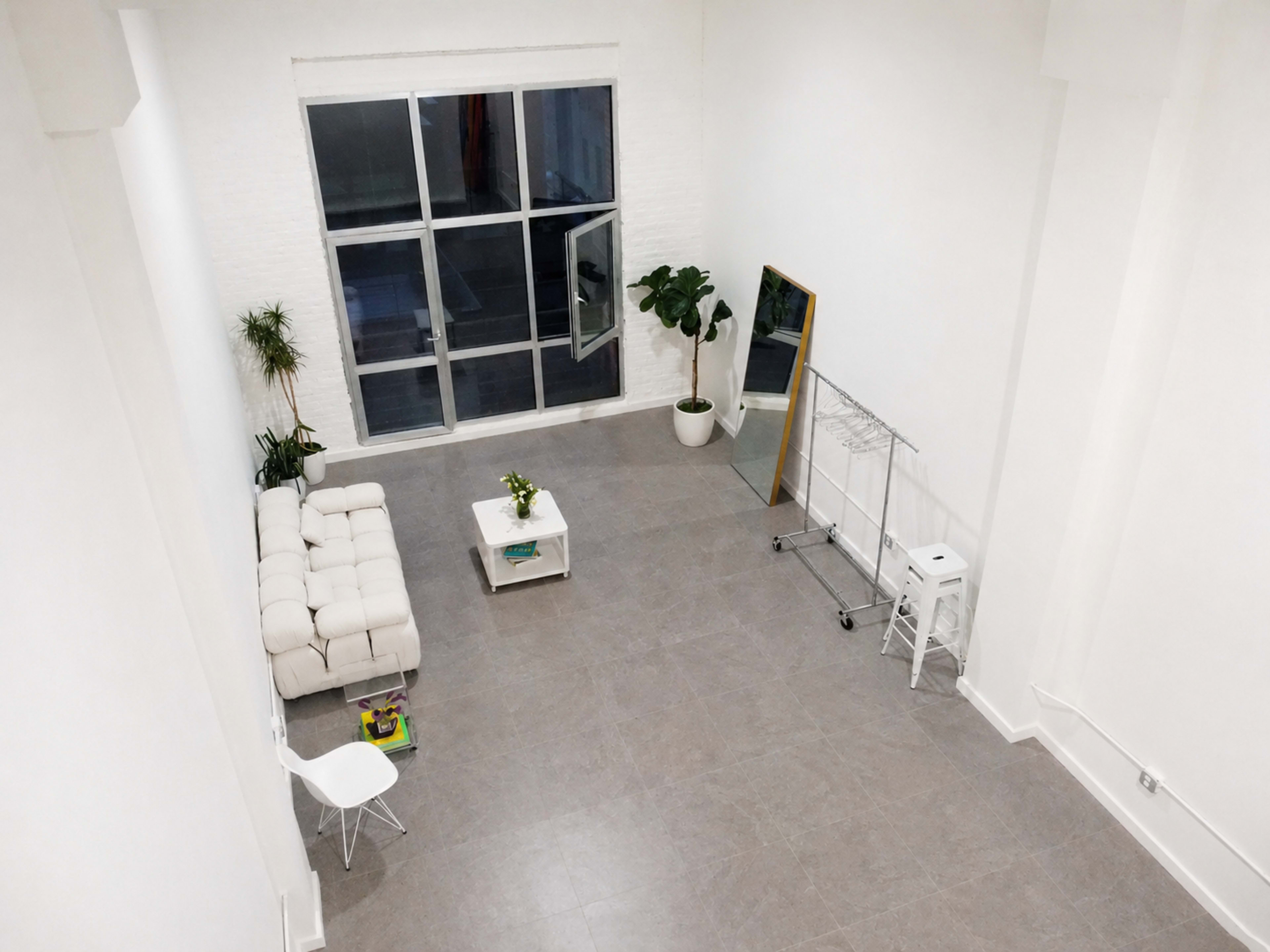 The image shows a spacious, minimalist room with a white sofa, a small coffee table, a potted plant, and a large window.