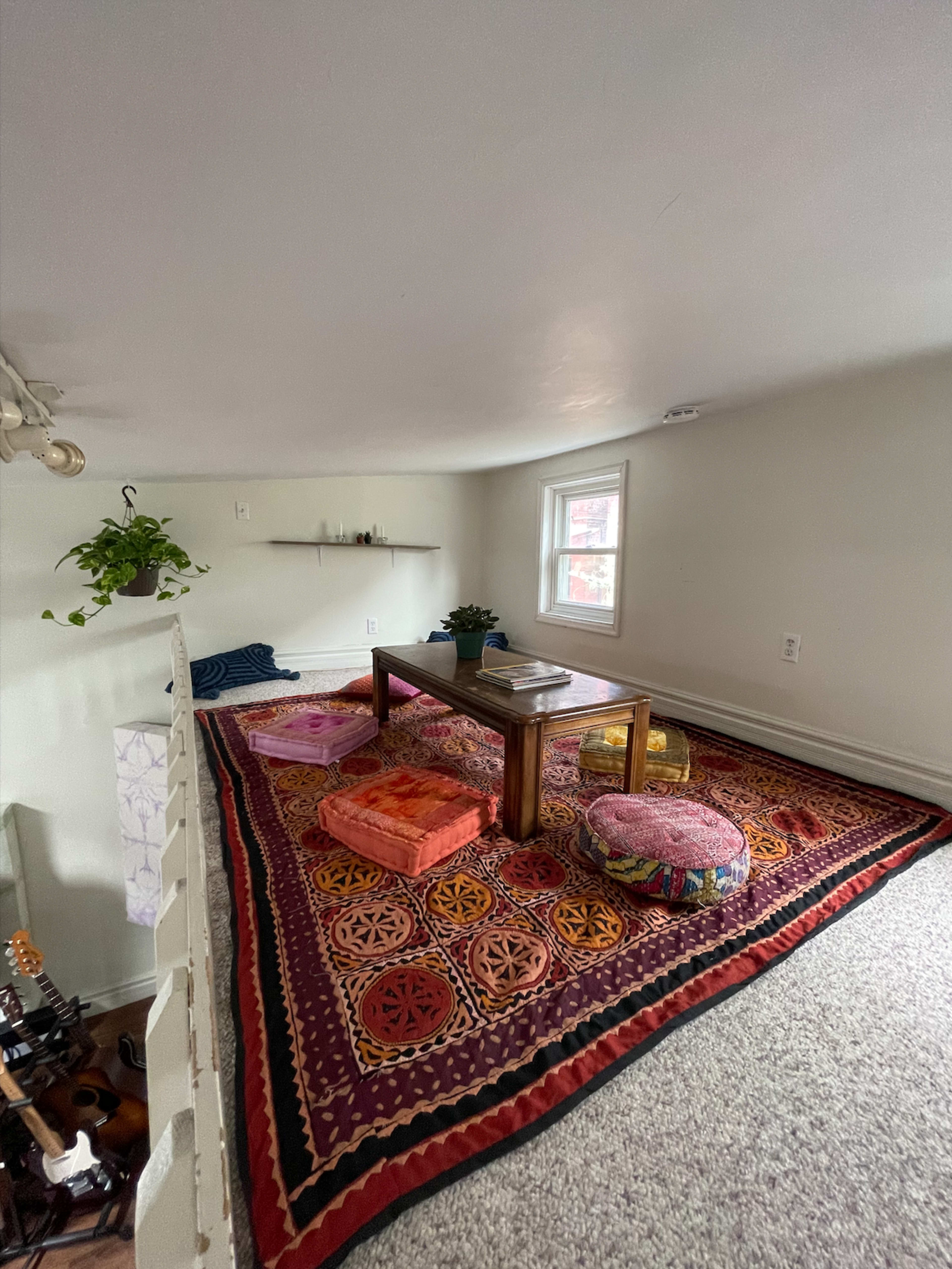 A small, cozy room with a patterned rug, a wooden table, and various cushions arranged for seating.