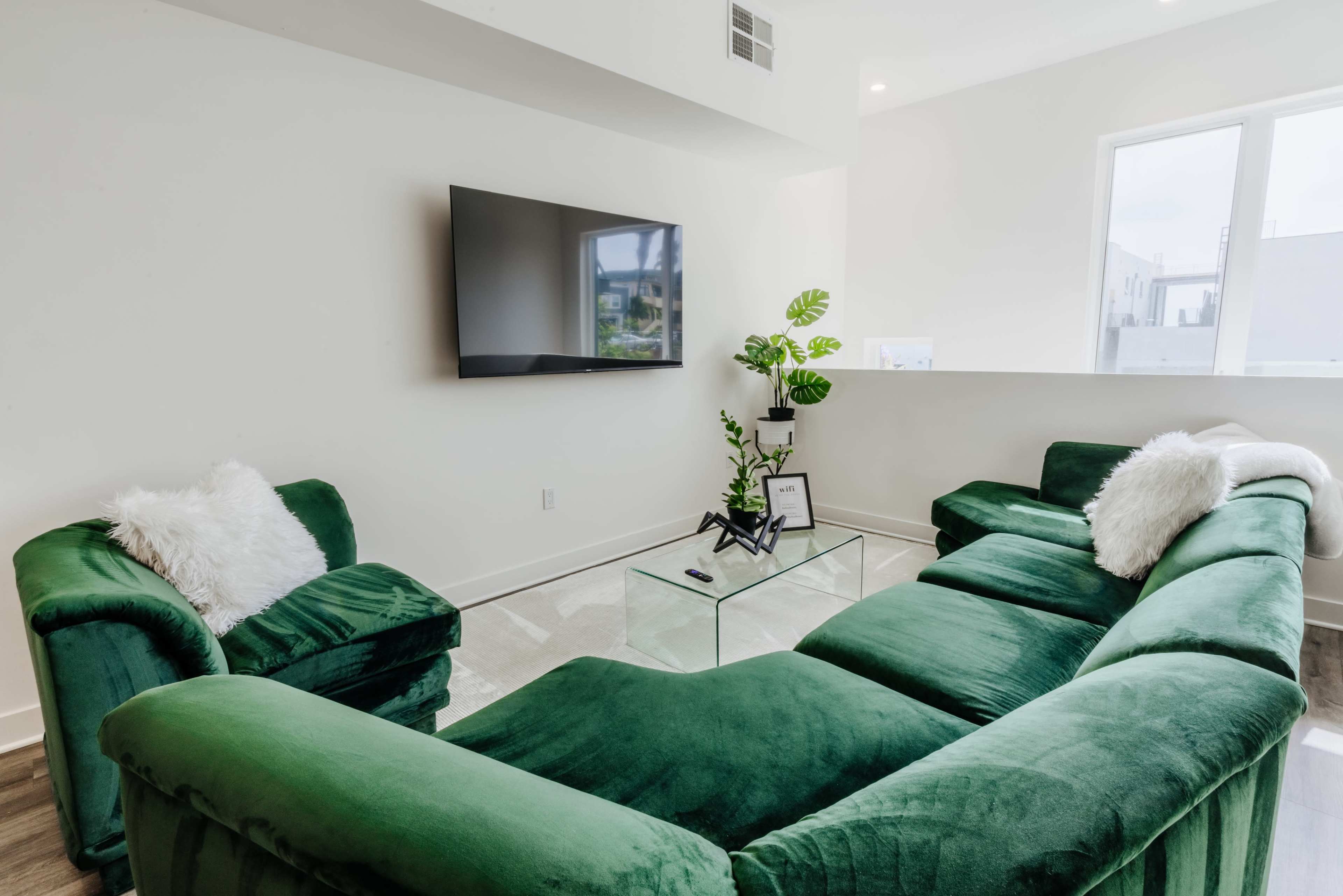 The living room features a green sectional sofa, two matching armchairs, a glass coffee table, and a television mounted on the wall.