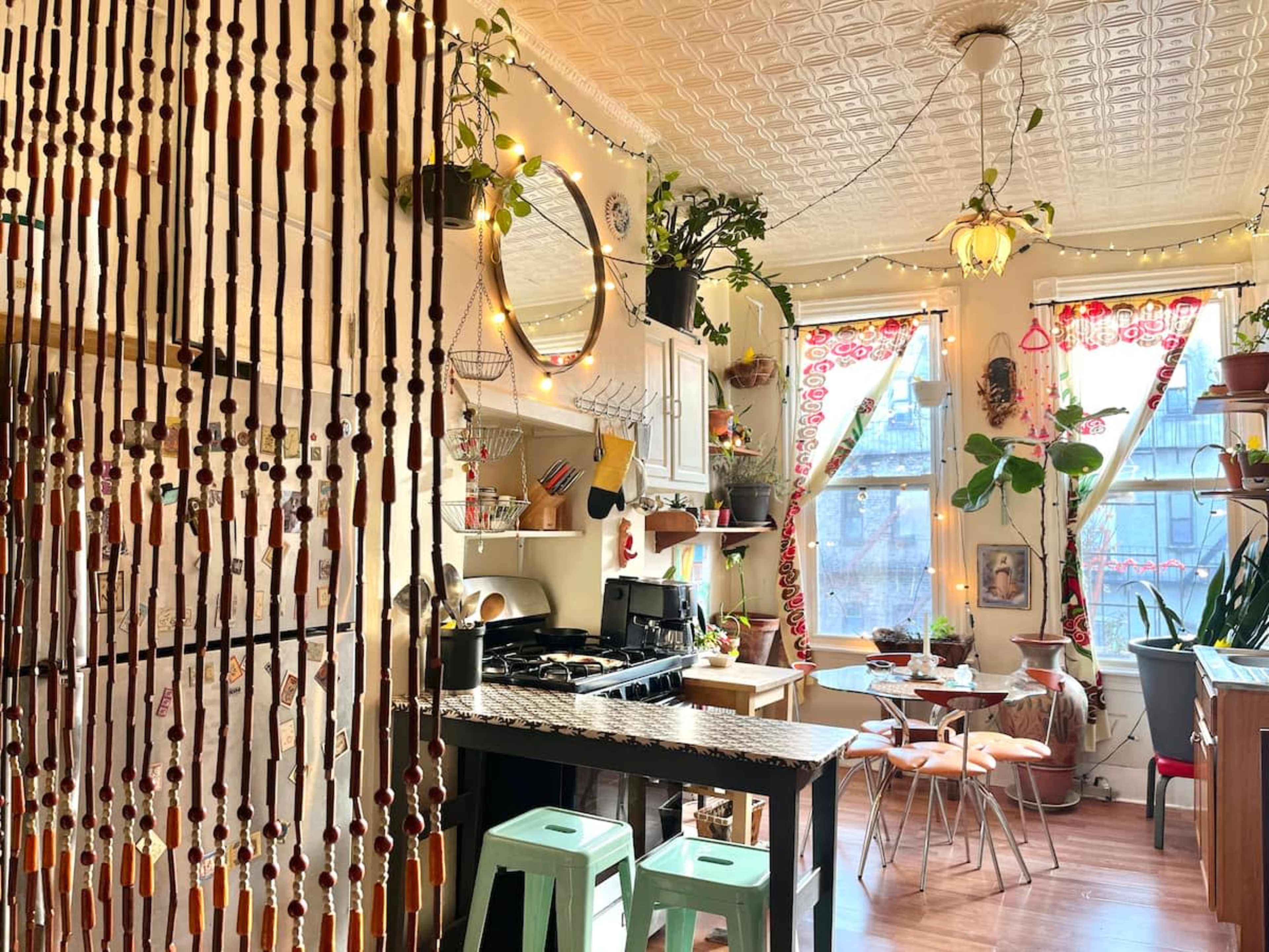 The image shows a cozy kitchen filled with plants, colorful decor, and a dining area with wooden furniture and bar stools.