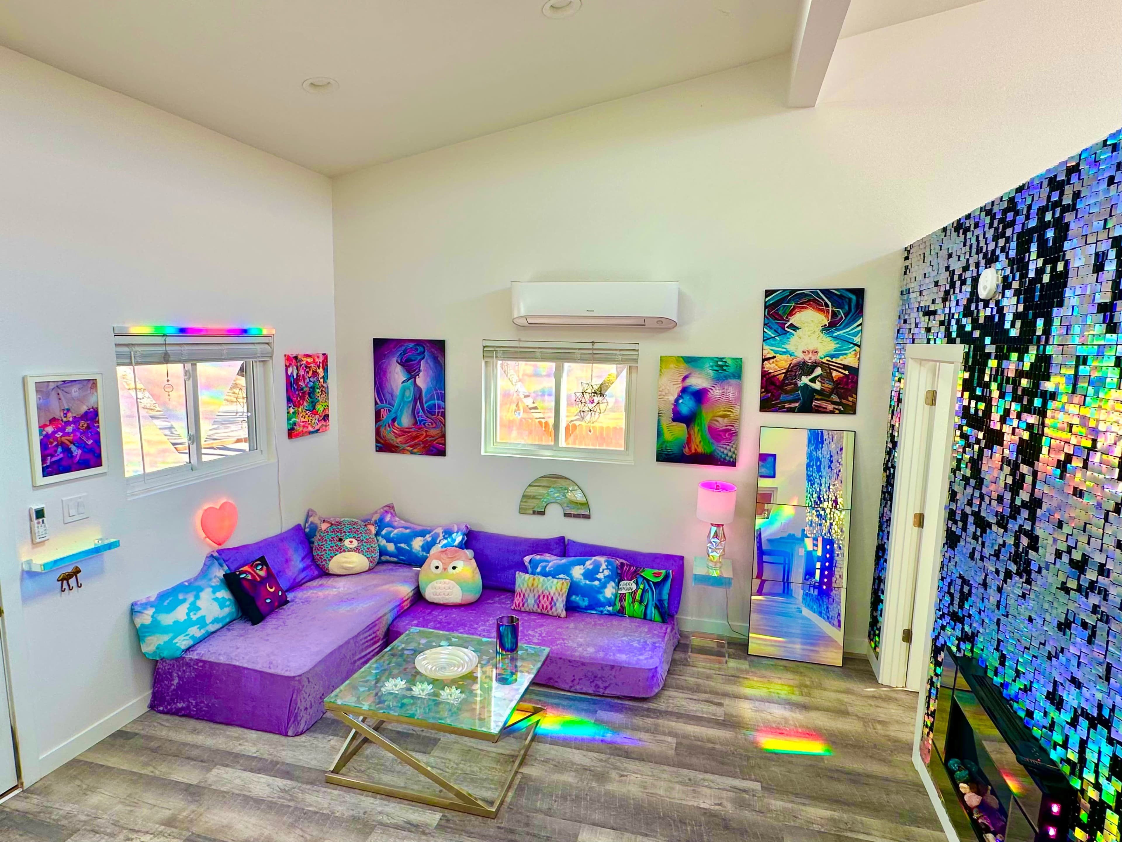 A brightly colored living room with modern furnishings, a large sectional sofa, decorative wall art, and a mirrored accent wall reflecting various colors.