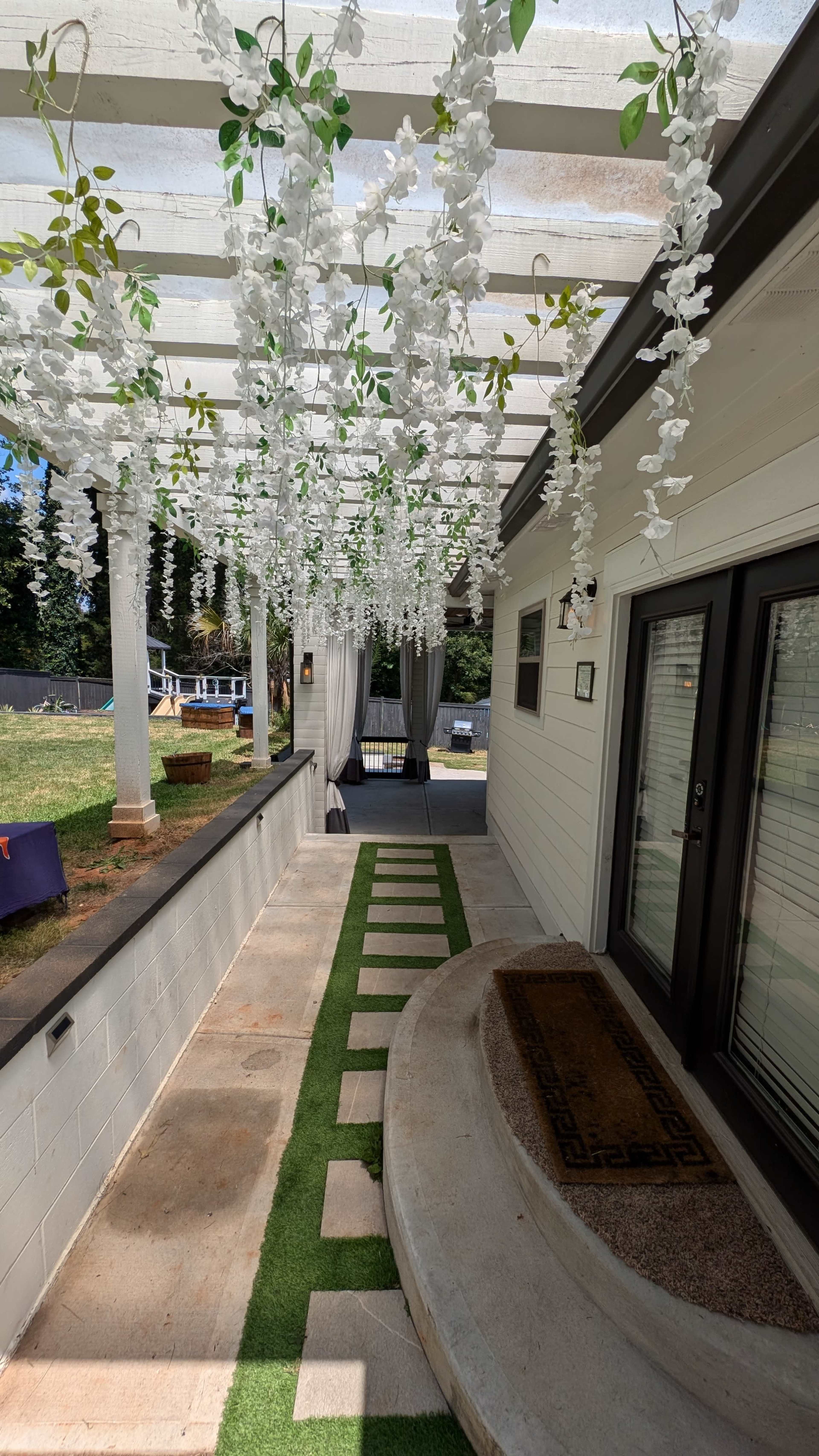 A pathway lined with artificial grass and stone stepping stones leading to a porch adorned with hanging white floral decorations.