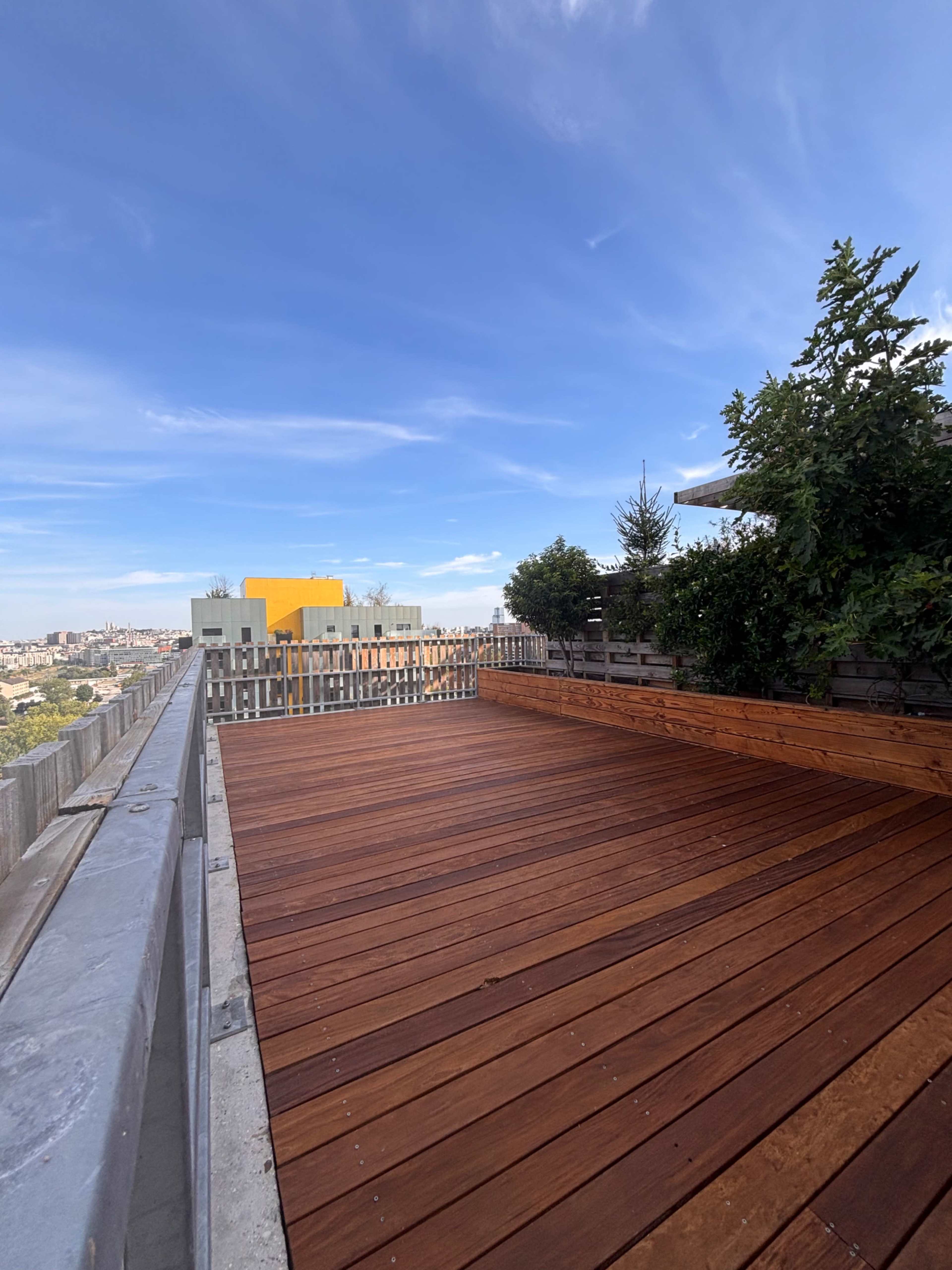 The image shows a wooden deck on a rooftop with a clear blue sky and some greenery along the edges.