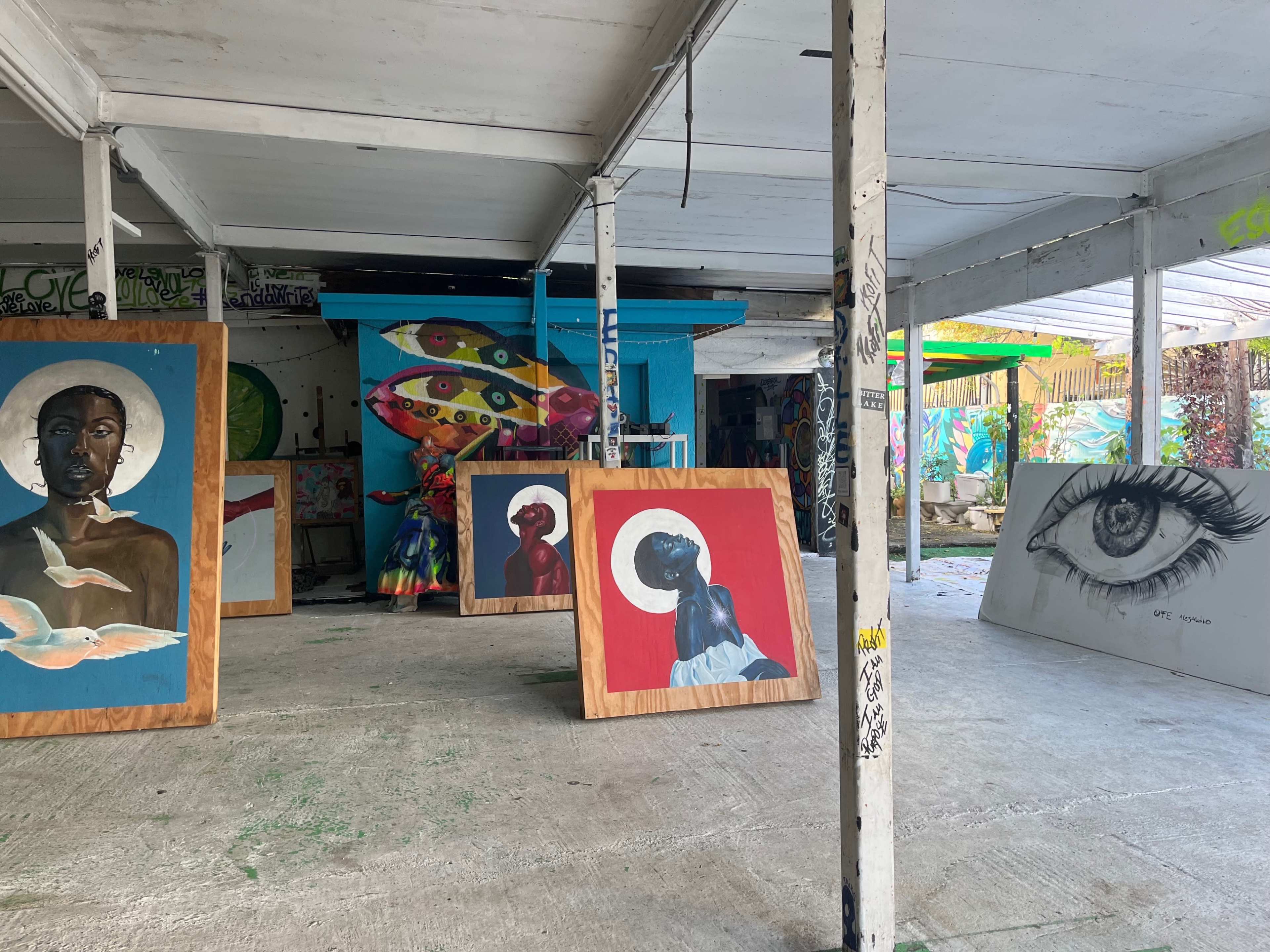 A gallery space with several large paintings displayed on the concrete floor, including portraits and a detailed eye sketch, set against vibrant murals on the walls.