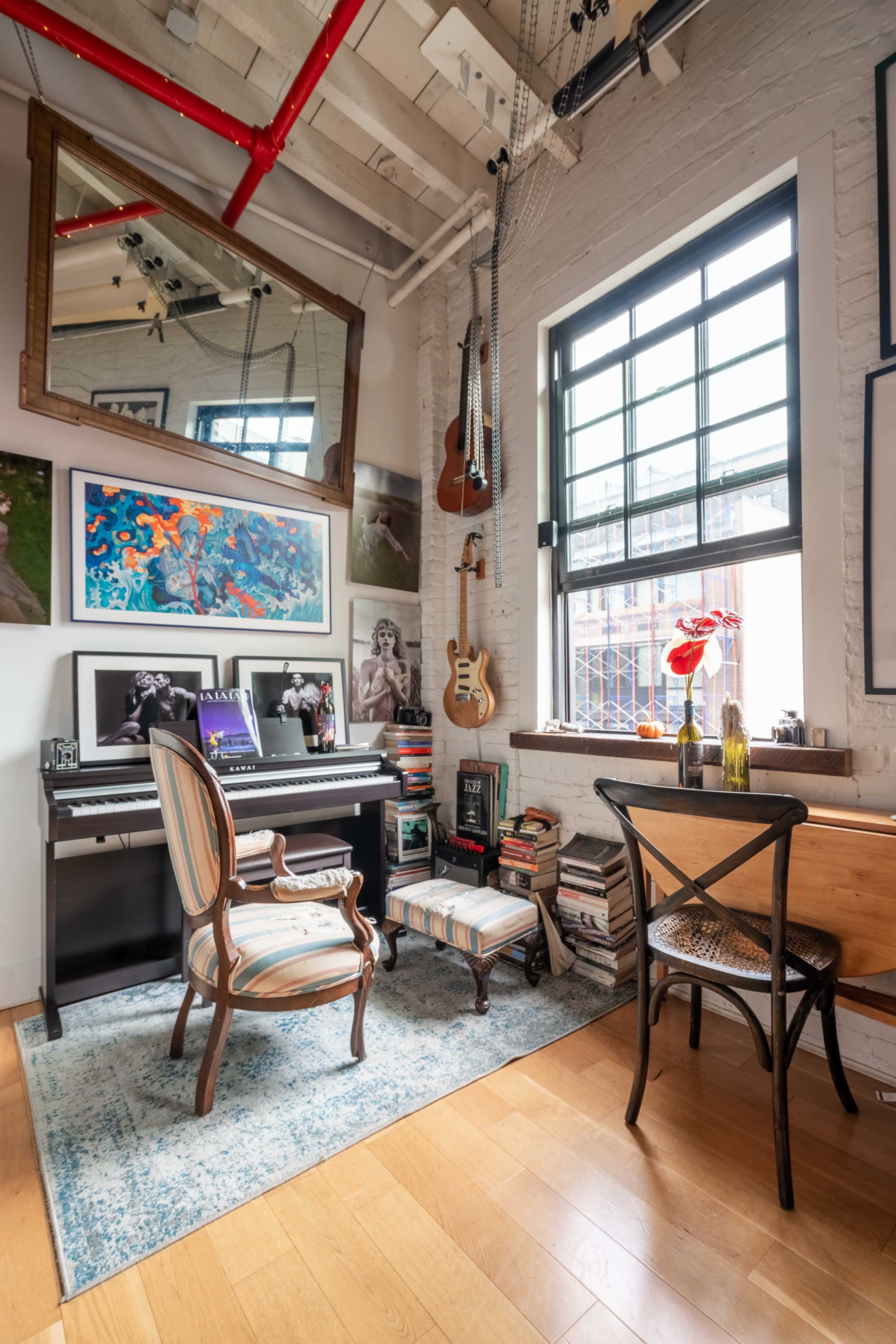 The image shows a cozy workspace featuring a piano, a chair, books stacked on the floor, and artwork displayed on the walls.