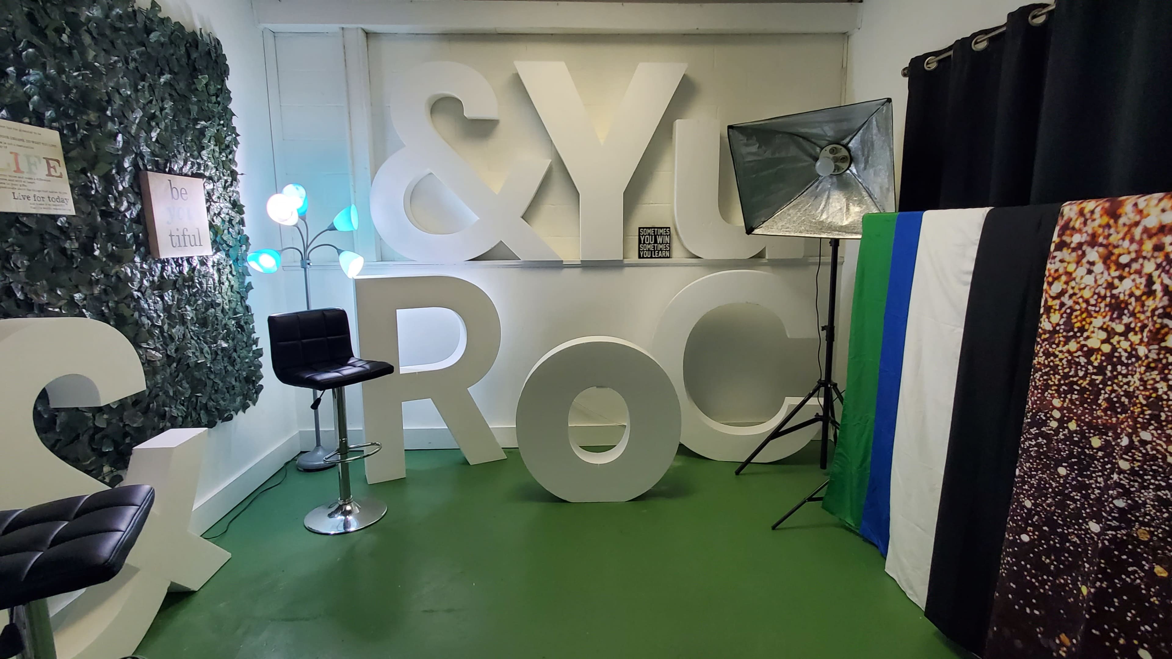 The image shows a room with large white letters spelling "& YOU ROCK," a black chair, a photography light setup, and a color-striped backdrop.