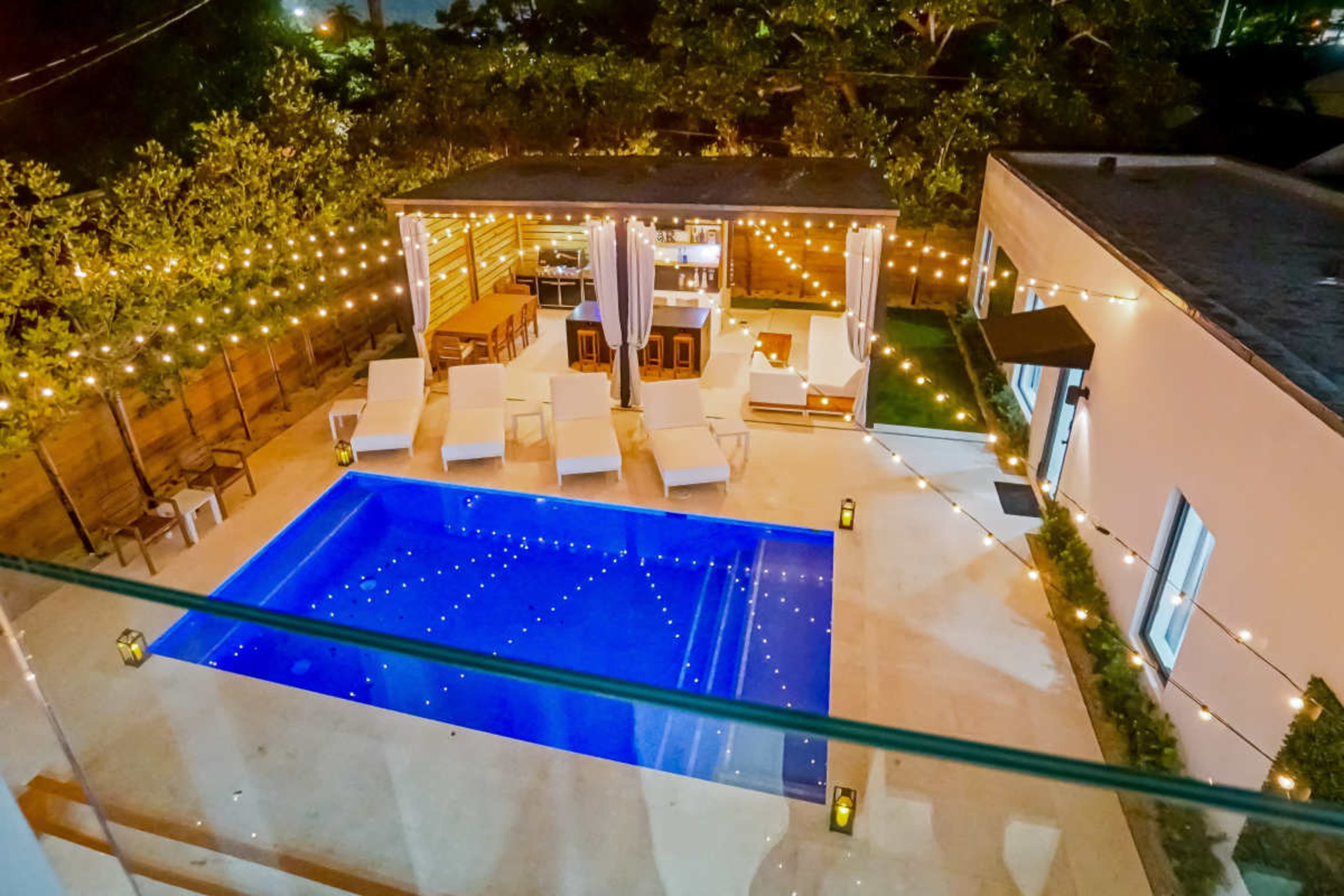 The image shows a well-lit outdoor pool area featuring a rectangular pool surrounded by lounge chairs, with a covered seating area and decorative string lights overhead.