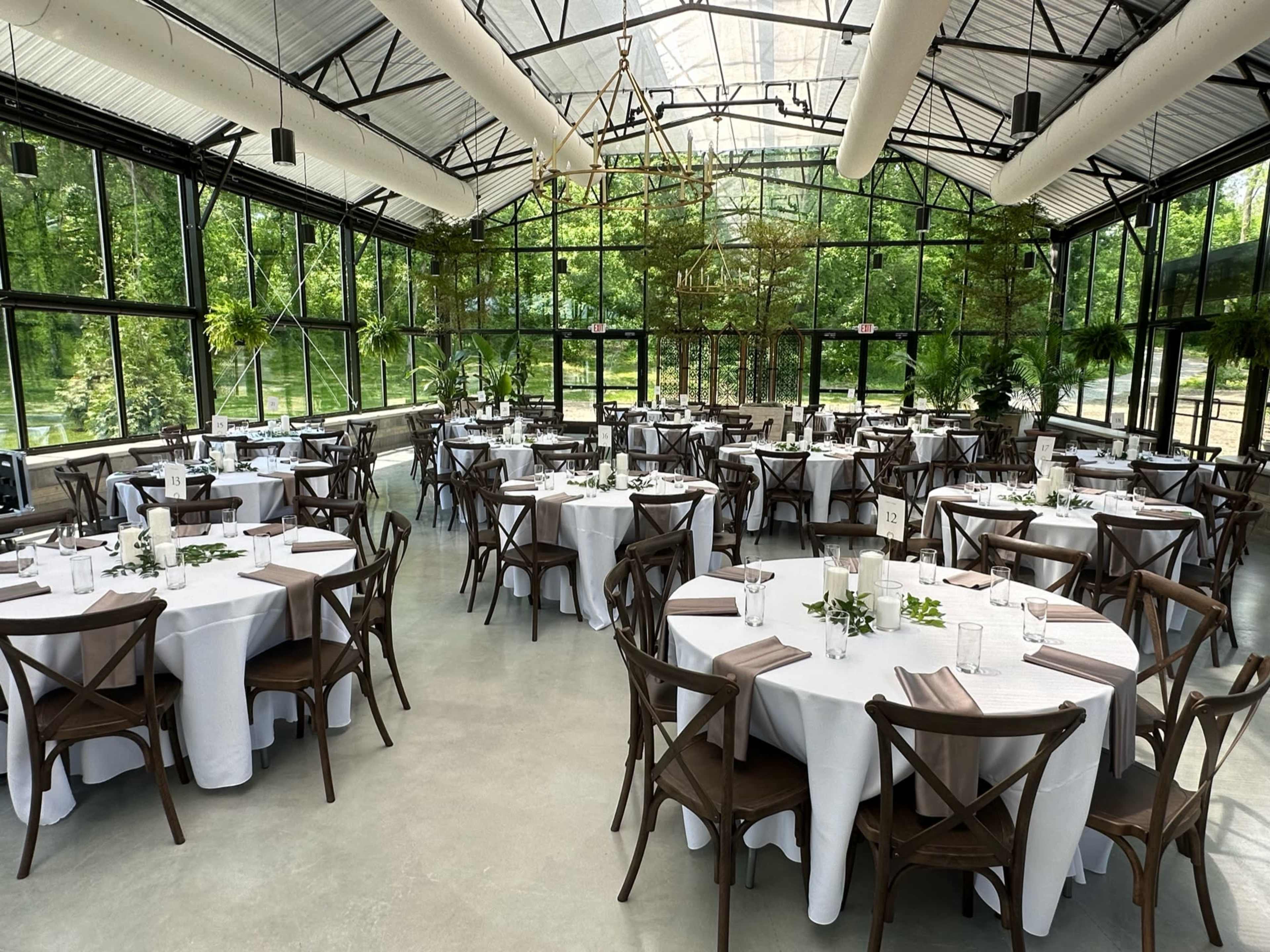 The image shows a spacious greenhouse venue set up for an event, featuring multiple round tables covered with white tablecloths and surrounded by wooden chairs.