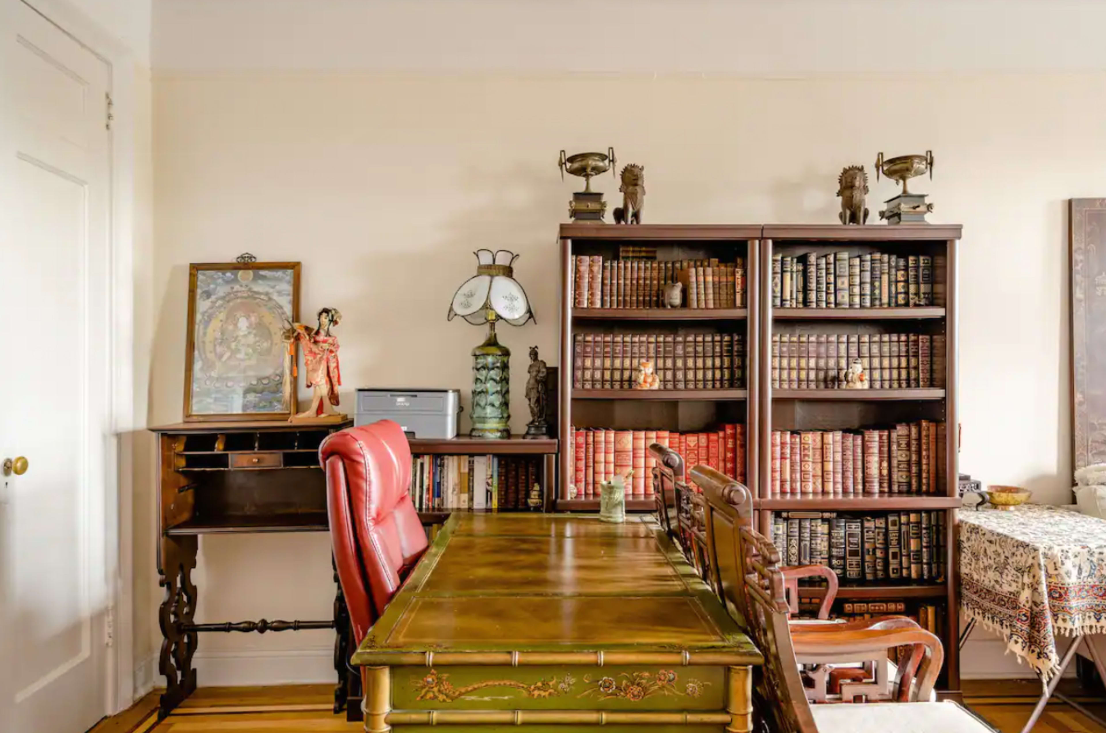 A vintage-style study with a wooden desk and red leather chairs facing a bookshelf filled with books, alongside a small writing desk and decorative items.