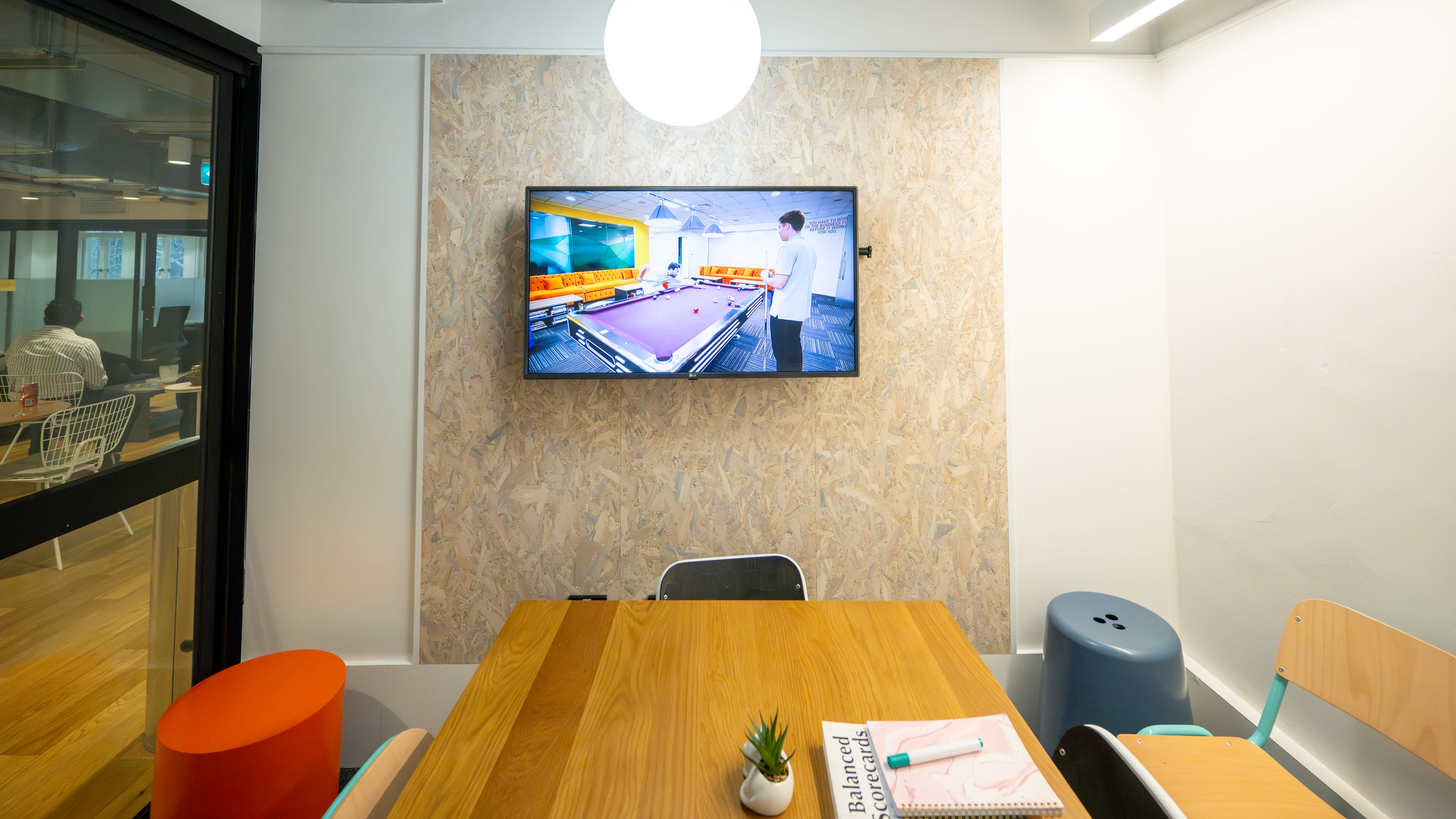 A modern meeting room with a wooden table, chairs in various colors, and a wall-mounted TV displaying a scene of a pool table.