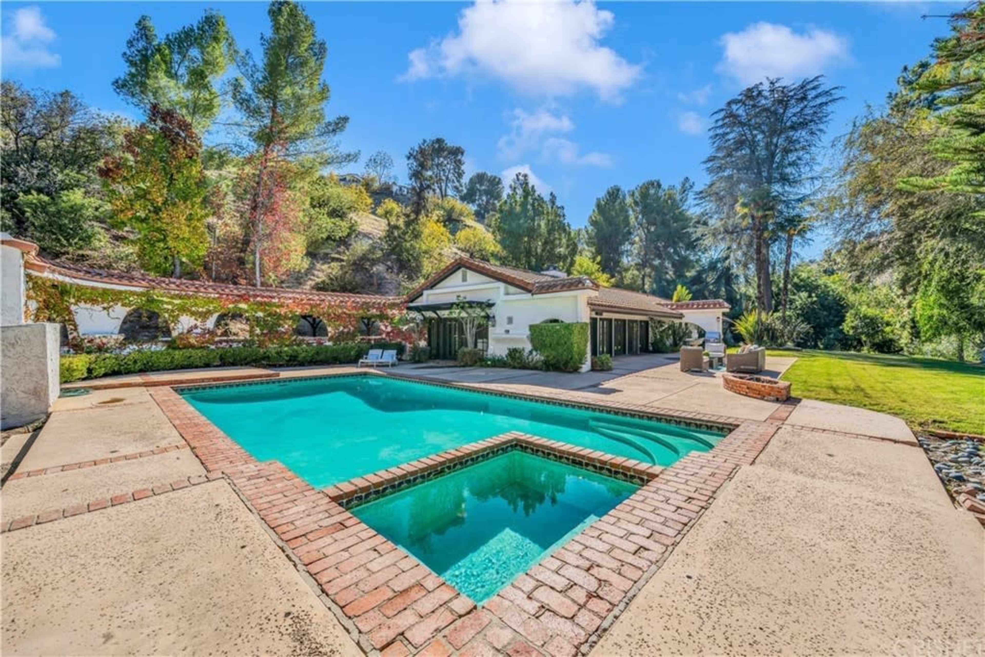 A pool with a brick patio is surrounded by green trees and a house with a tiled roof.