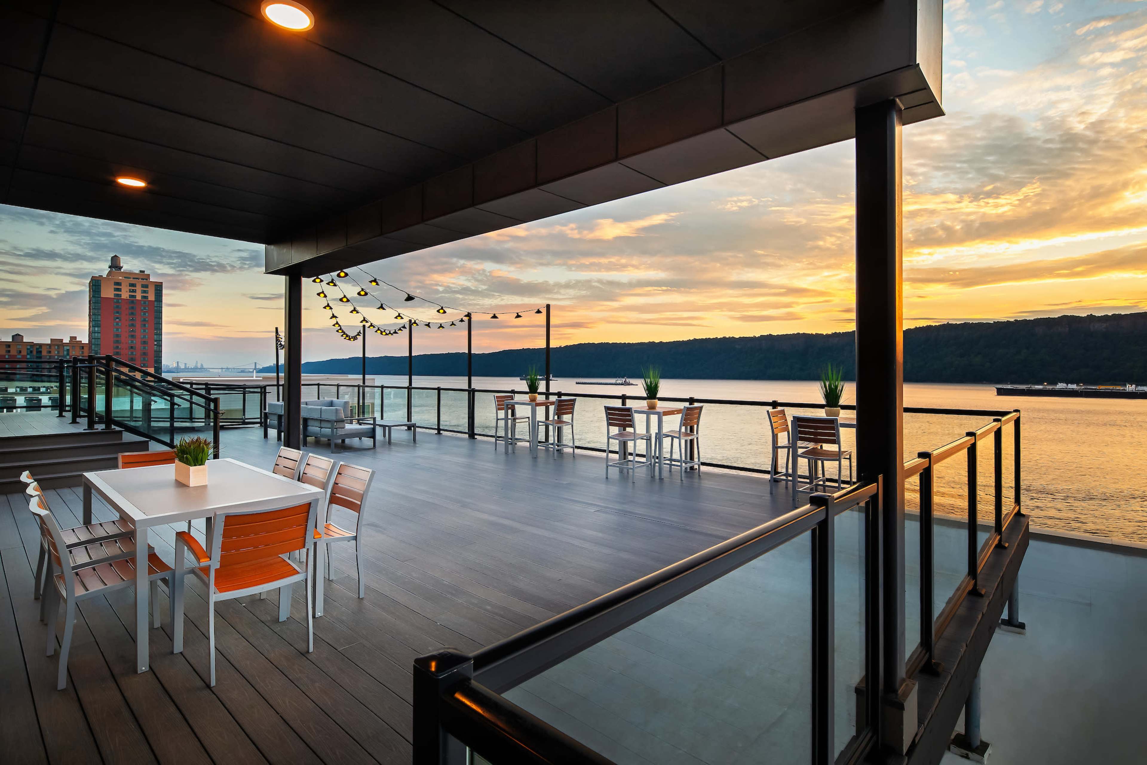 The image shows a modern outdoor deck with tables and chairs overlooking a river at sunset.