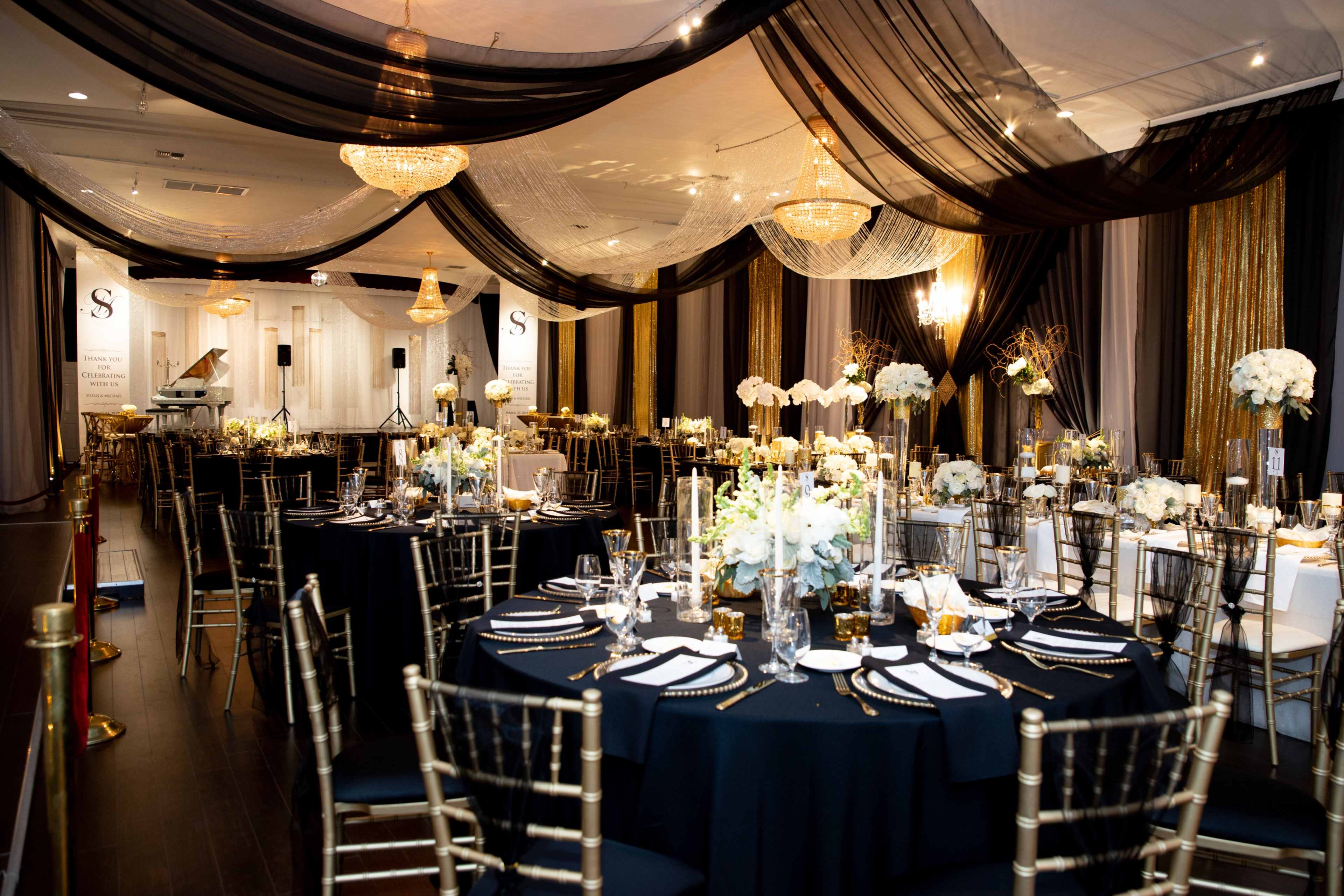 The scene depicts an elegantly decorated event space with round tables set for a formal dinner, adorned with white floral arrangements and gold accents.