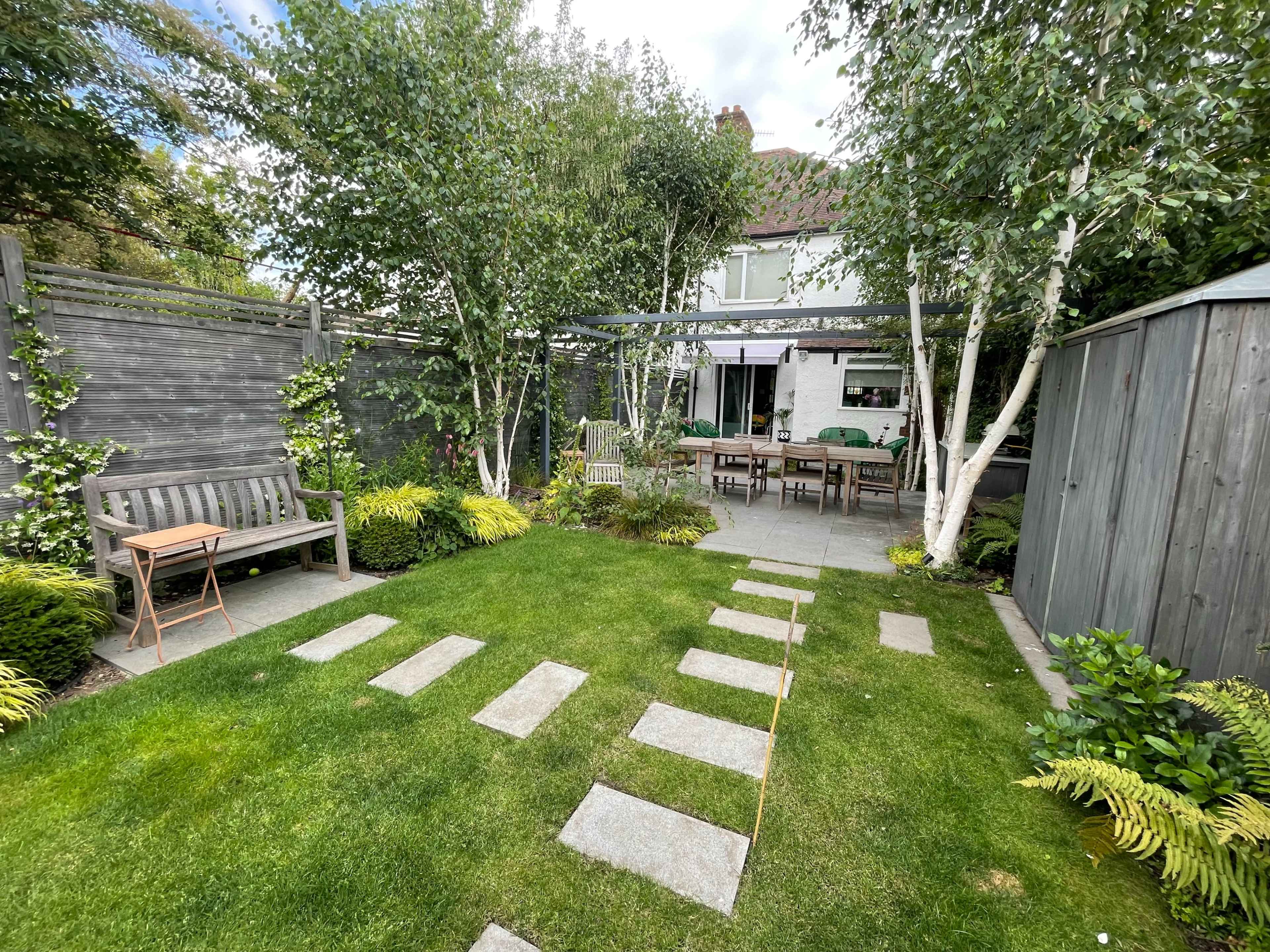 A landscaped garden with stone pathways, greenery, and seating areas, leading to a dining table under a pergola.