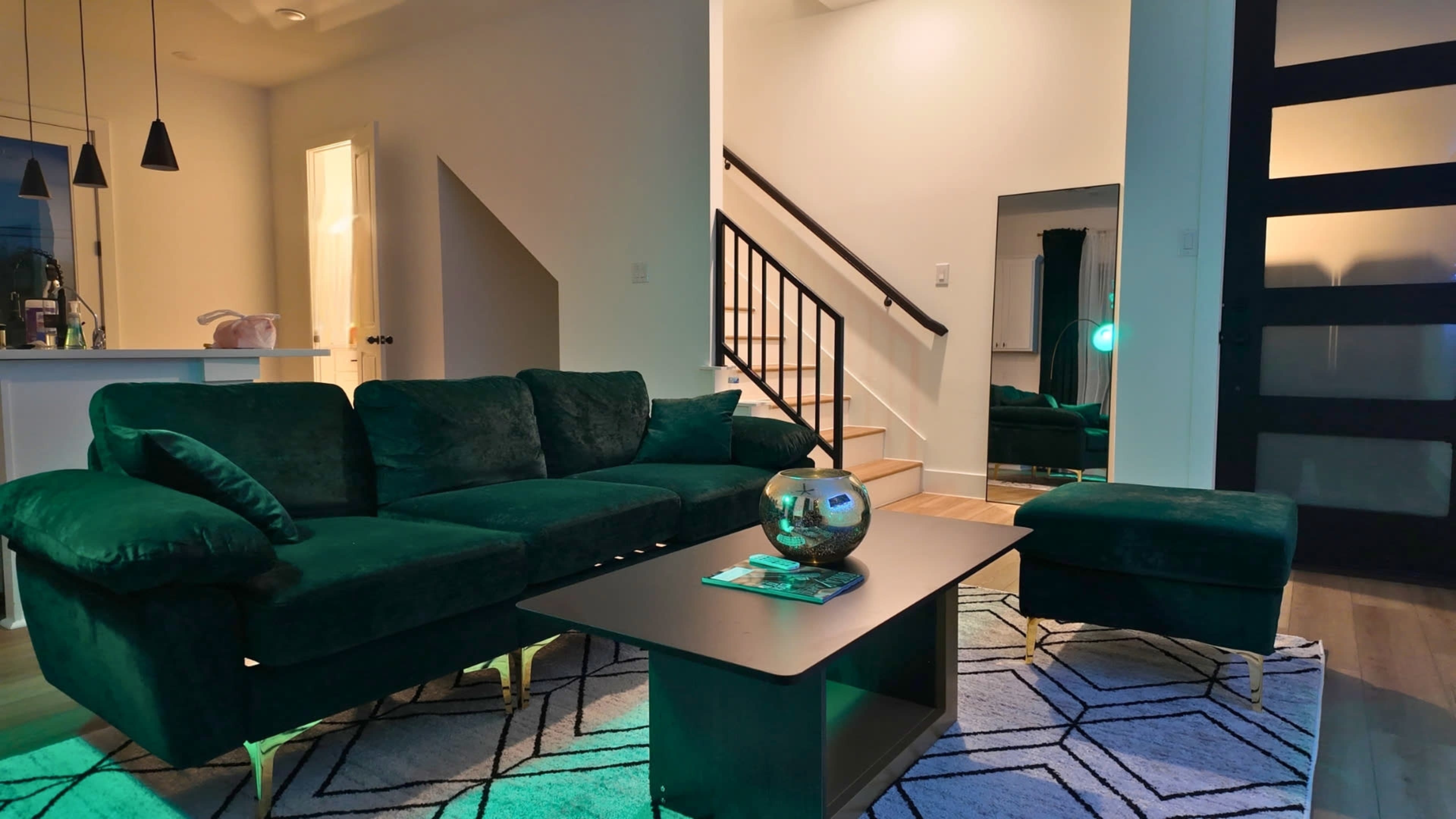 The image features a modern living room with a green velvet sofa, a geometric patterned rug, a coffee table, and a staircase in the background.