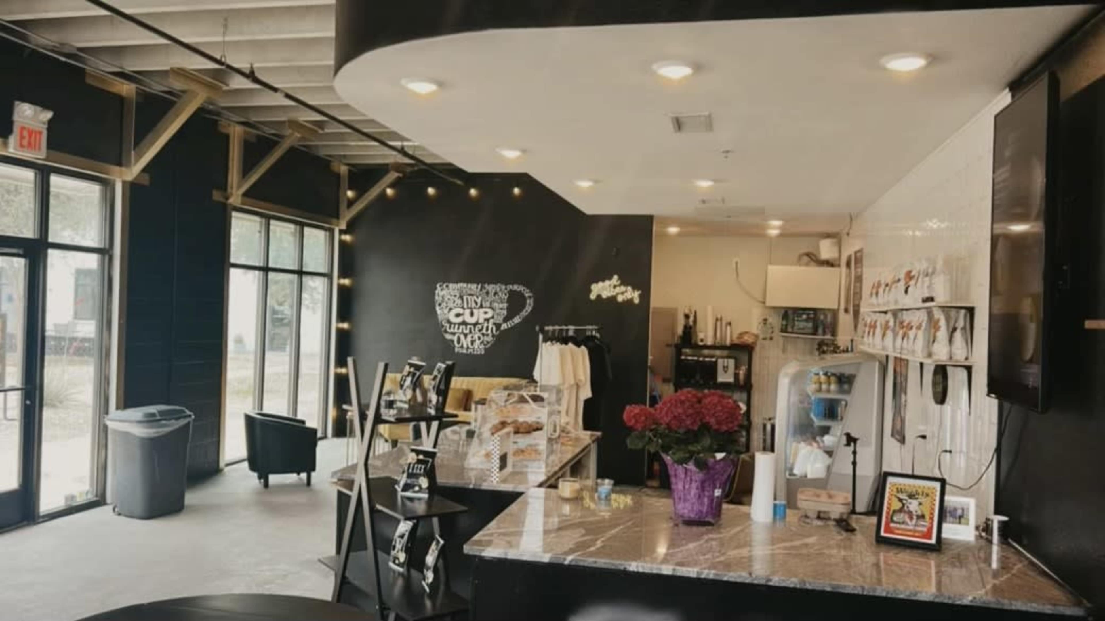 A modern café interior features a counter with baked goods, a seating area with black tables, and large windows allowing natural light to enter.