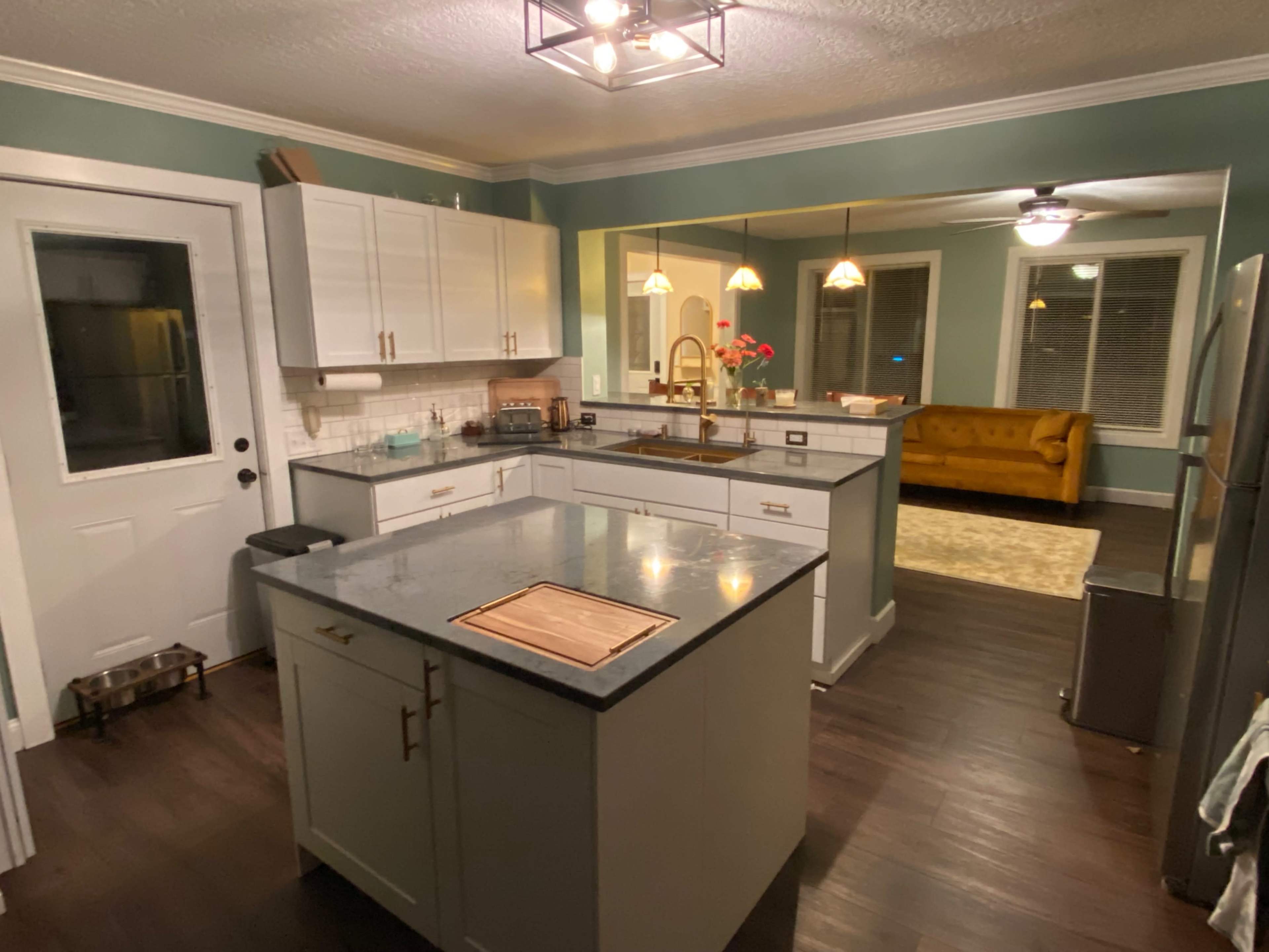 The image shows a modern kitchen featuring white cabinets, a large island with a gray countertop, and a cozy living area with a yellow couch visible through a doorway.